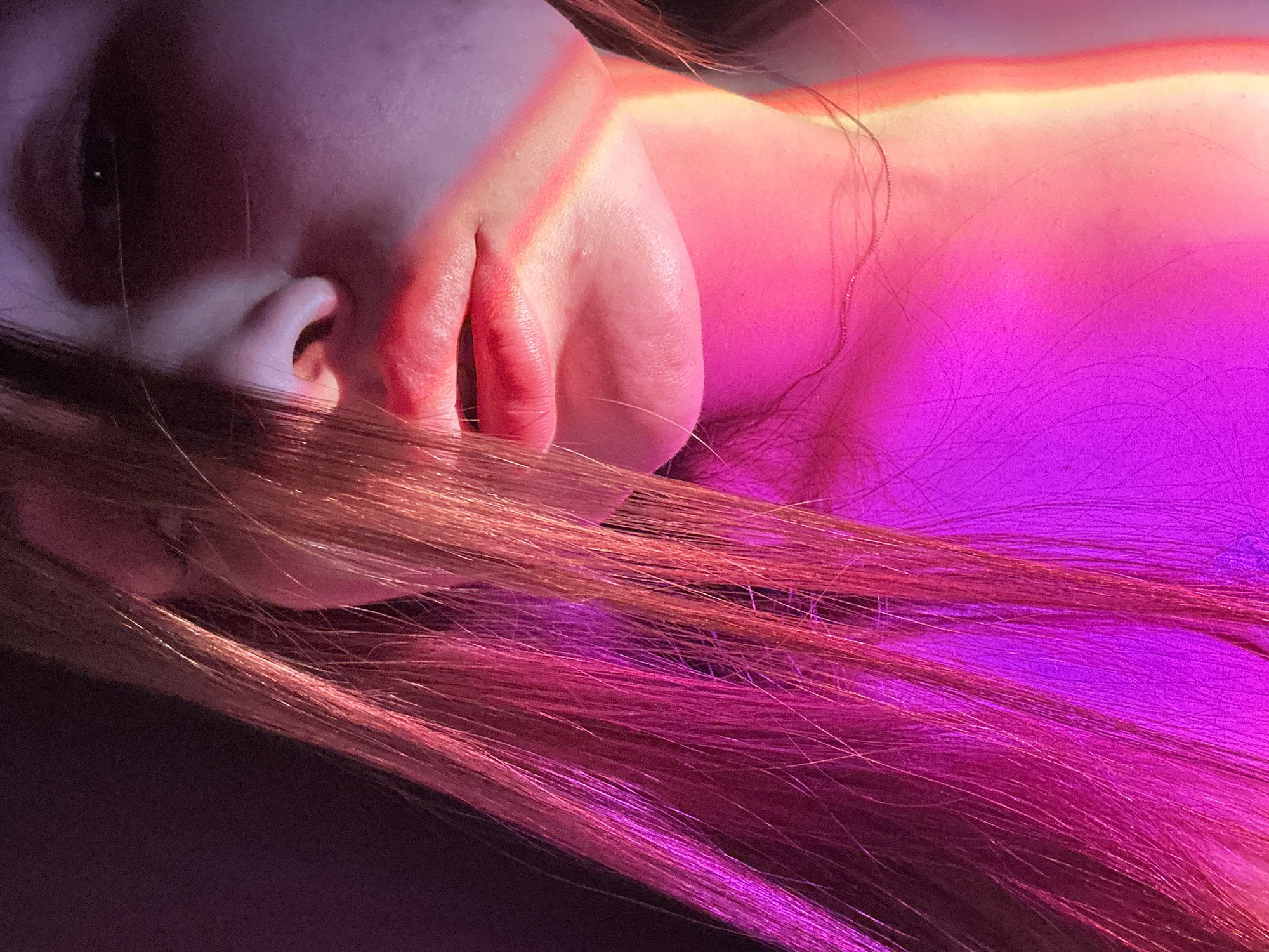 A close-up shot presents a person's face and chest area illuminated with vivid, artificial lighting, predominantly in shades of pink and purple, with hints of orange or yellow on the upper chest. The person is lying down, with their head tilted slightly, and strands of light brown hair fall across their face. The visible eye is dark, and the facial features are somewhat obscured by shadows and hair. The skin appears fair, and the lips are slightly parted. There is no indication of the location being in the city of Chaikovsky, Russia, within the visual information provided. The background is dark and indistinct.