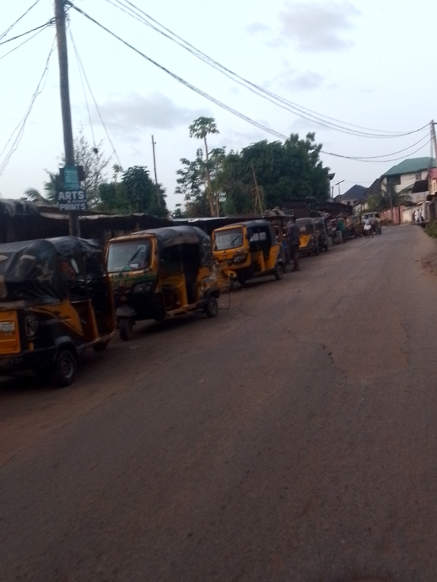 The image shows a row of auto rickshaws, also known as "keke" or "tricycles," parked along a paved street in what appears to be a city. The rickshaws are predominantly yellow and black. A sign reading "ARTS & PRINTS" is visible near a utility pole. Trees and buildings line the background. Several people are visible further down the street. The location is Unknown, Unknown.