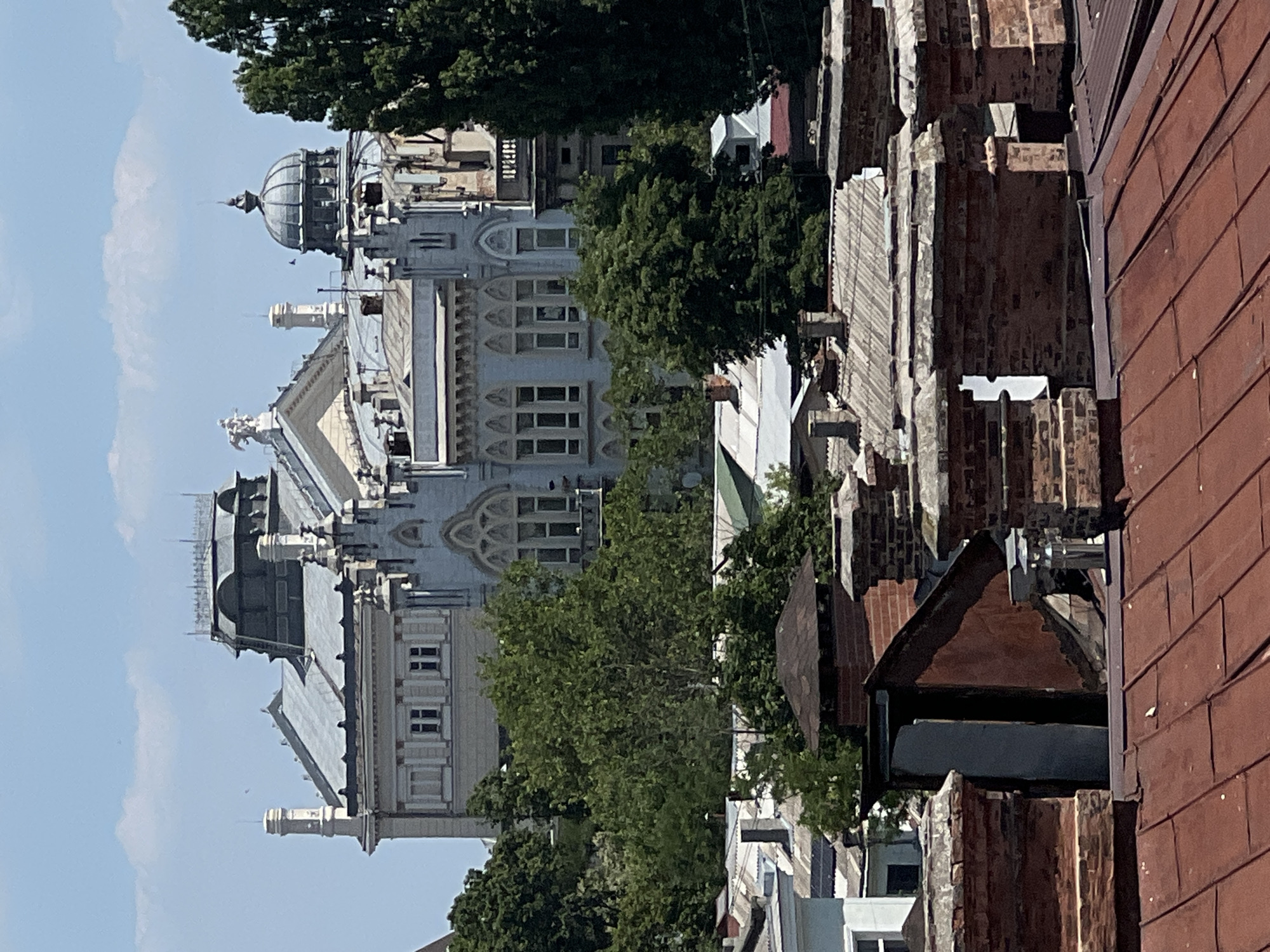 This media file shows a view of Odesa, Ukraine, from a rooftop perspective. The scene is dominated by the architecture of older buildings, characterized by ornate facades, domes, and intricate window designs. In the foreground, a reddish-brown tiled roof is visible, with a section of unfinished brickwork and a dark, angular structure. Beyond this, a large, pale gray building with multiple levels and arched windows takes center stage. It features a prominent dome on its left side. Lush green trees are interspersed throughout the scene, partially obscuring some of the structures and adding a natural element to the urban landscape. The sky is a clear, bright blue, suggesting a sunny day, likely midday given the lighting. There are no people or overt activities depicted; the focus is on the static architectural elements and the surrounding environment. The overall impression is one of historical depth and a preserved urban aesthetic under a pleasant sky.