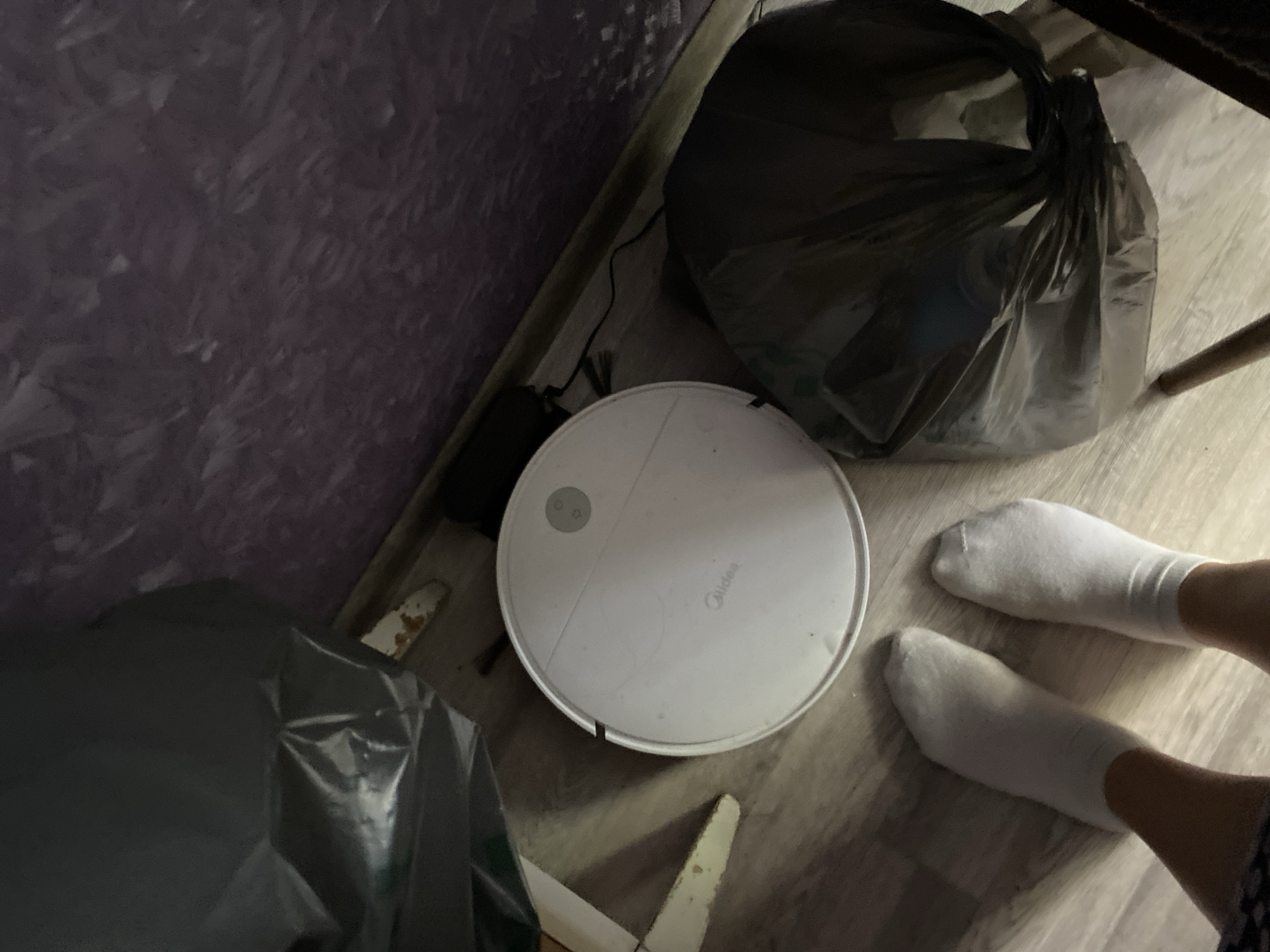 The image depicts a low-angle interior shot of a room, likely a domestic setting, with a focus on a white robot vacuum cleaner positioned on a light-colored wooden floor. Two white socks are visible in the foreground, suggesting a person's feet are present but not fully in frame. The robot vacuum has the "Midea" logo on its surface. To the left and right of the vacuum, two large black trash bags are present, one partially obscuring the vacuum cleaner. In the background, a textured purple wall is visible, and a black power cord snakes across the floor. There are no visible signs or indications linking this scene to the city of Lomonosov, Russia.