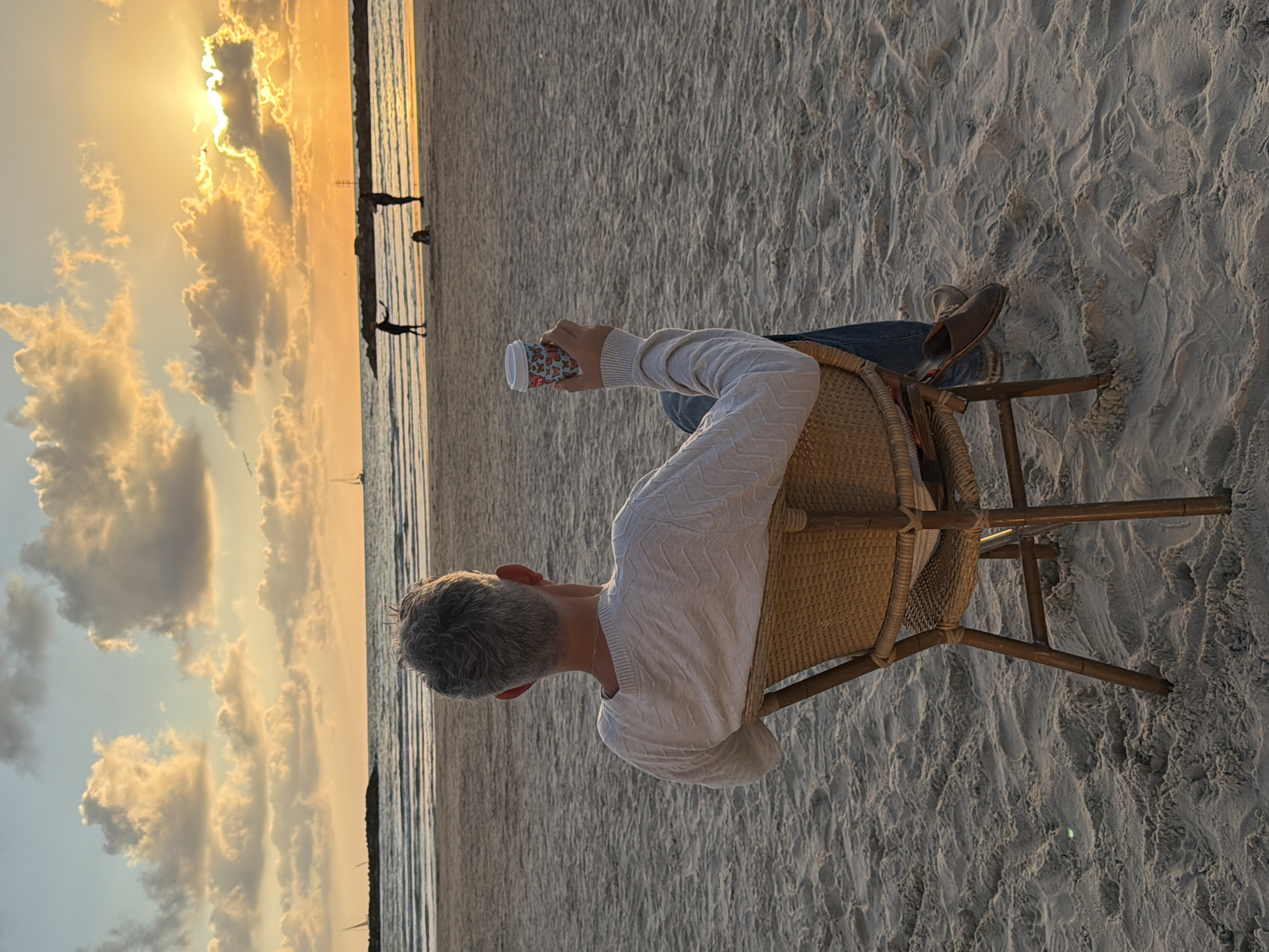 A person with gray hair sits in a wicker chair on a sandy beach facing the Mediterranean Sea at sunset. The person wears a white sweater and blue jeans and holds a patterned cup. People are silhouetted in the distance along the shoreline. The location is likely near Tel Aviv, Israel, as indicated by the Mediterranean coastline. The beach is sandy, and the sky is filled with clouds reflecting the setting sun.