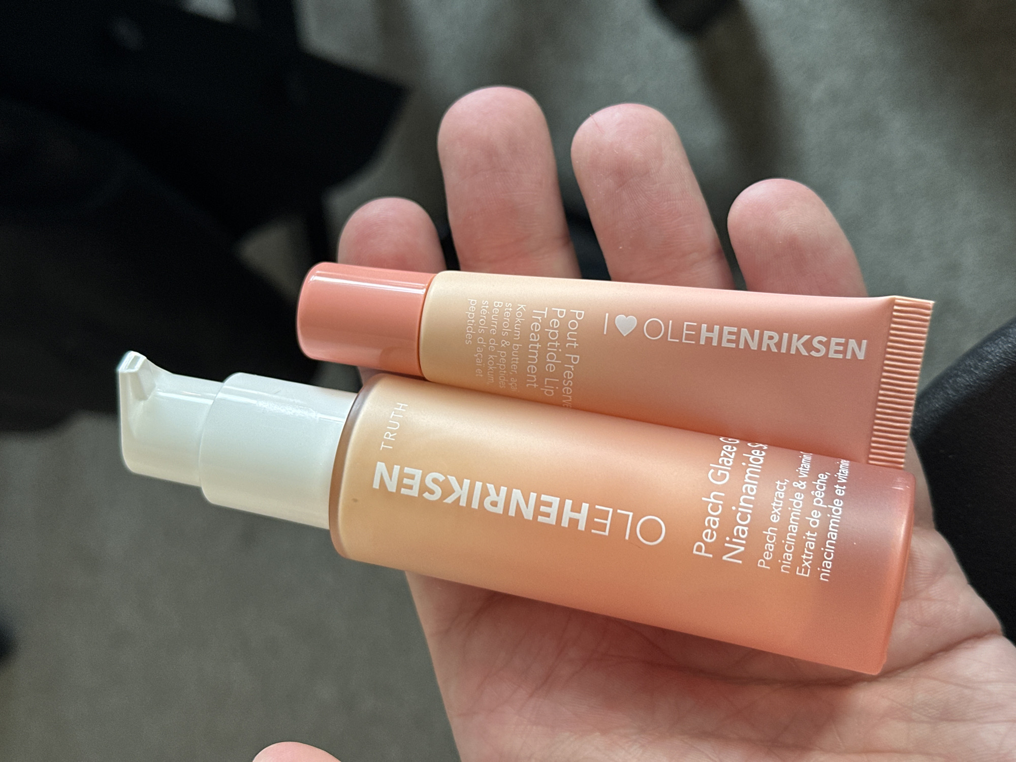 A person's left hand holds two peach-colored OLEHENRIKSEN skincare products. The larger product on the left is an upright bottle with a white pump dispenser, labeled "OLEHENRIKSEN TRUTH Peach Glaze Niacinamide Serum," indicating peach extract, niacinamide, and vitamin content in English and French. The smaller product on the right is a tube, positioned slightly lower than the bottle, labeled "Pout Preserve Peptide Lip Treatment," detailing kokum butter, açaí sterols, and peptides as ingredients in English and French, and displaying "I love OLEHENRIKSEN." The background is out of focus, consisting of dark grey and black tones, suggesting an indoor setting. The specific location in London, United Kingdom, cannot be determined from the visual evidence provided in the image.