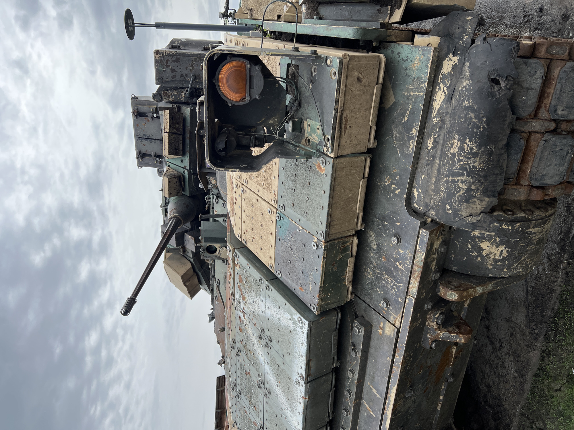 The image shows a close-up view of the front of a military tank. The tank is heavily armored with bolted metal plates. The left side of the tank is visible and covered in dirt. A gun barrel protrudes from the turret. On the front of the tank, there is a damaged light fixture with an orange bulb. The background shows a cloudy sky. There are no people visible. The location is not ascertainable from the image.
