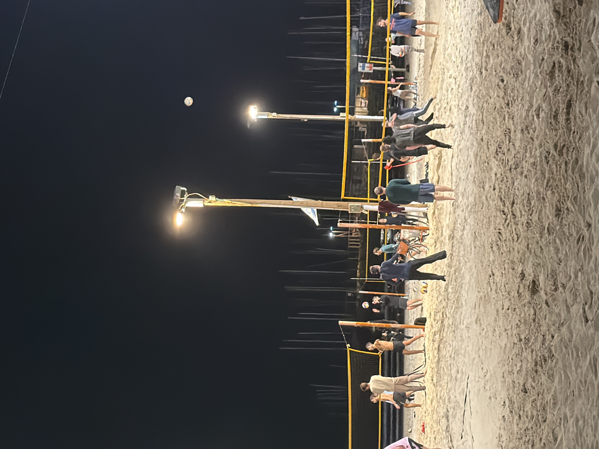 At night, a beach volleyball game takes place on the sandy beach in Tel-Aviv, Israel. A volleyball is suspended in the air above the net. Several groups of people are standing on the beach. The area is illuminated by overhead lights.