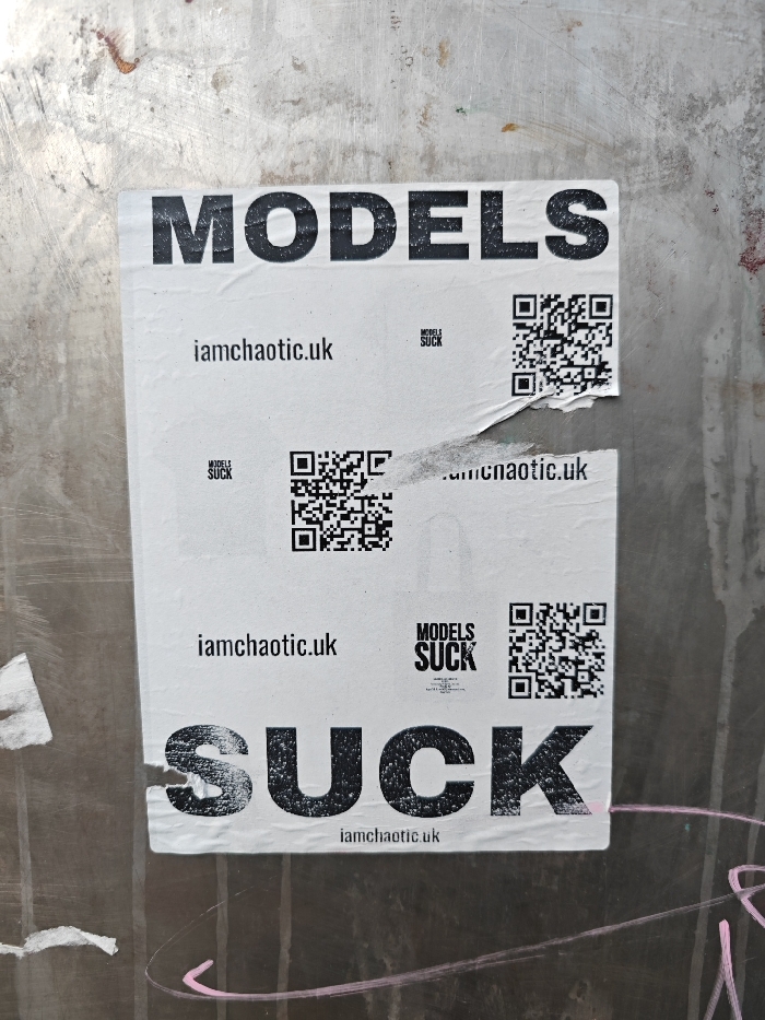 A white paper poster, measuring approximately A2 size, is affixed to a textured, light grey metallic or concrete surface in an urban environment, specifically in London, United Kingdom. The poster features large, sans-serif black text, with "MODELS" prominently displayed at the top and "SUCK" at the bottom. The body of the poster contains repeated black text entries of "iamchaotic.uk" and "MODELS SUCK" in smaller font sizes, each accompanied by a distinct QR code. Three full or partial instances of this pattern are visible: one below "MODELS", one partially torn horizontally in the middle right section, and one above "SUCK". The poster exhibits signs of wear, including wrinkles, creases, and multiple tears, particularly the central horizontal tear. The background surface is discolored with grime, subtle reddish-brown stains, and faint pink chalk or paint marks in the lower right corner.