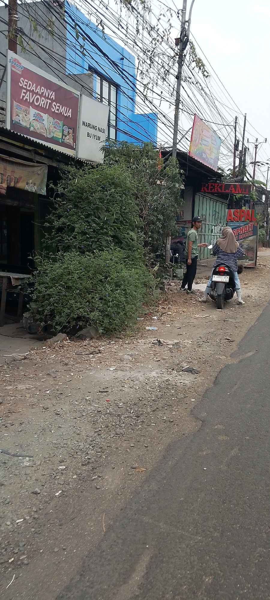 A low-angle, outdoor, full shot captures a street scene with two individuals and a motorcycle in Bekasi, Indonesia. The foreground shows a dirt and gravel shoulder next to an asphalt road. In the midground, a green bush is prominent on the left. To the right of the bush, a man wearing a green t-shirt and black pants stands near a motorcycle. A woman, wearing a beige hijab and a striped shirt, is seated on the motorcycle. Behind them, a storefront with a corrugated metal roof and a light blue building facade is visible. Several signs are present: one on the left reads "SEDAPNYA FAVORIT SEMUA," another in the center reads "WARUNG NASI BU IYUM," and a red LED sign on the right displays "REKLAMI." A sign for "ASPAL" is also visible on the right. The environment appears somewhat unkempt, with debris on the ground. Electrical wires are strung across the scene overhead.
