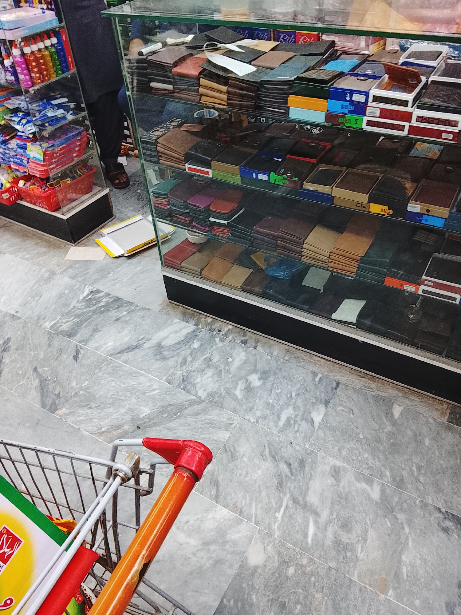 The image shows the interior of a shop in Gulgasht Colony, Pakistan. A glass display case contains stacked wallets and boxes of wallets. A person wearing dark clothing and sandals stands beside the case. A shopping cart with a red handle is in the foreground. The floor is tiled with gray marble. There are items on a shelf to the left of the display case. A discarded yellow and white cardboard box sits on the floor between the shelf and the display case.