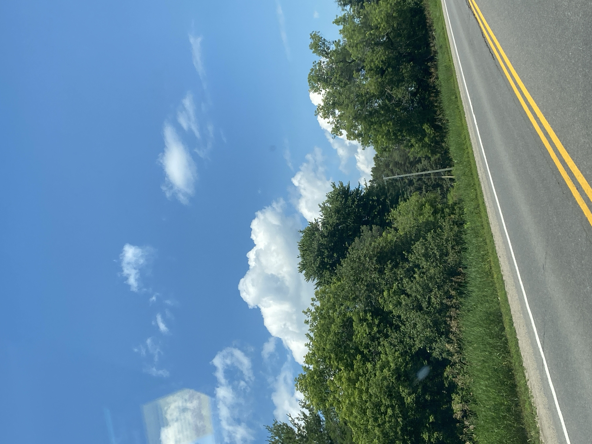 The image depicts a road with double yellow lines and a white line on the right side, bordered by a grassy shoulder and a dense line of green trees. The sky above is a clear blue with scattered white clouds. A utility pole is visible in the background among the trees. The scene is likely captured from a moving vehicle, suggesting a journey through a rural or semi-rural area. The image does not contain any people or specific structures that would pinpoint a precise location within Saint-André-Avellin, Canada, but it evokes a typical roadside landscape found in the region during daytime.