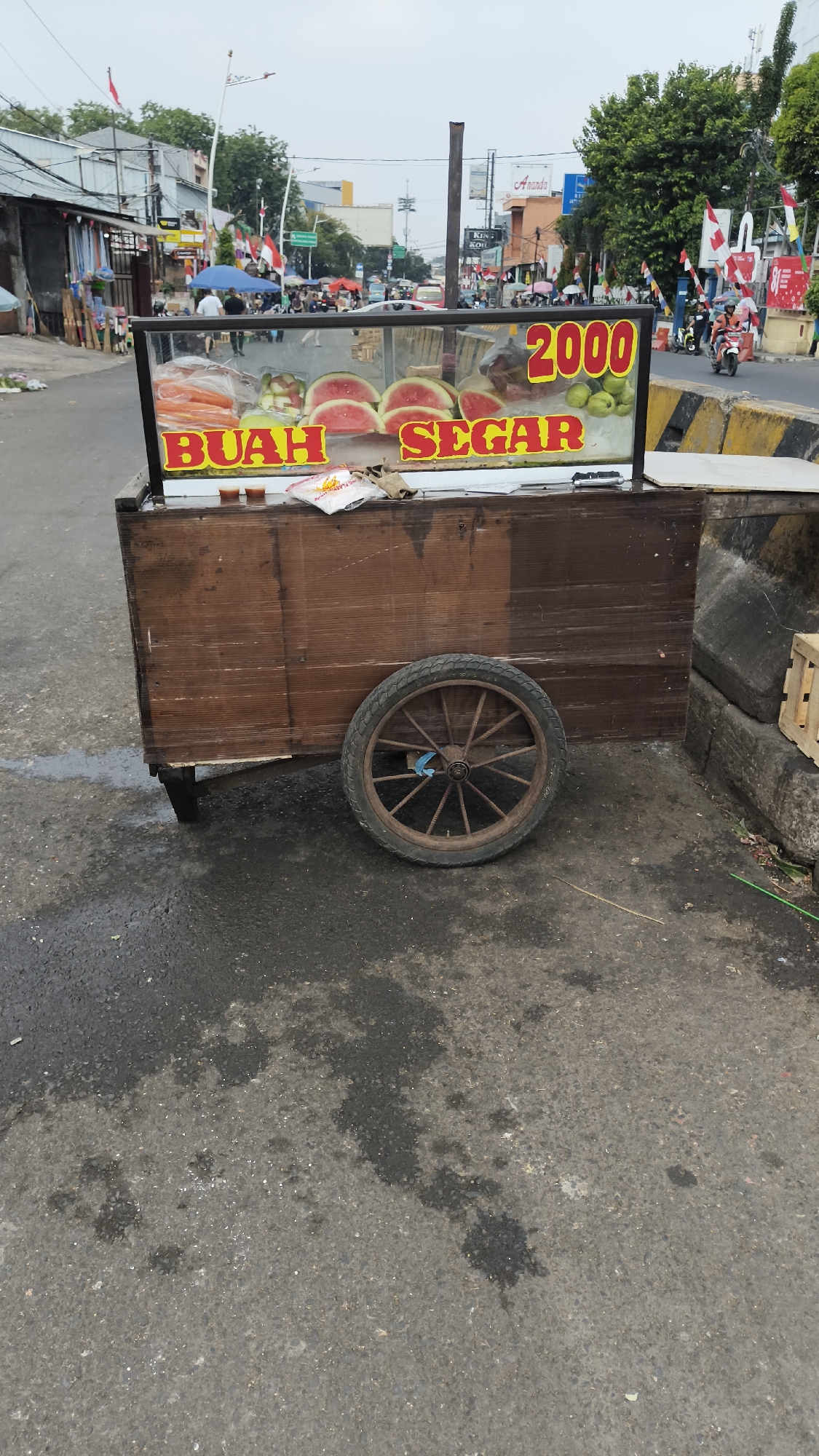A fruit cart is parked on the side of a busy street in South Jakarta, Indonesia. The cart is made of wood and has a glass display case filled with sliced watermelons and other fruits. A price of "2000" is visible on the display. The street is paved with asphalt and shows signs of wear and tear, with water stains and debris scattered on the surface. In the background, there are several shops and buildings, as well as people walking and riding motorcycles. The sky is overcast, suggesting a cloudy day.