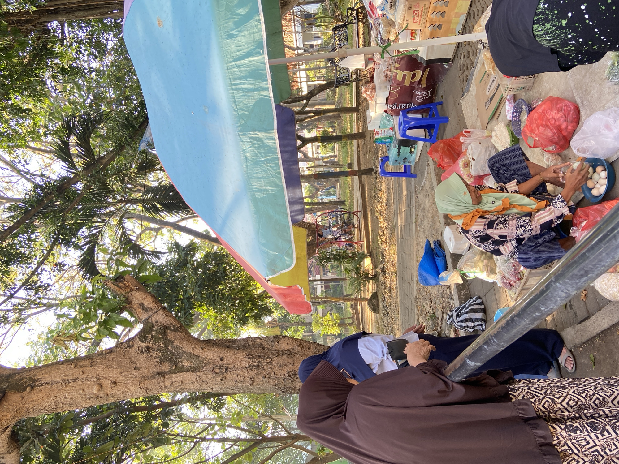 In a marketplace setting in Kecamatan Pati, Indonesia, a street vendor wearing a light green hijab and patterned clothing is crouched down, arranging eggs in a blue bowl. She is surrounded by various goods, including red plastic bags filled with produce, white plastic bags, and baskets of items. To her right, another person, wearing a dark hijab and a black top with white speckles, is partially visible, also facing the stall. In the background, a large, colorful umbrella provides shade over the stall. Two women in hijabs are standing to the left, seemingly interacting with the vendor or observing the marketplace. The ground is paved with stone, and trees with lush green foliage create a natural backdrop. Several blue plastic stools and boxes are also present, along with a bicycle in the distance. The scene is illuminated by natural sunlight, suggesting it is daytime.