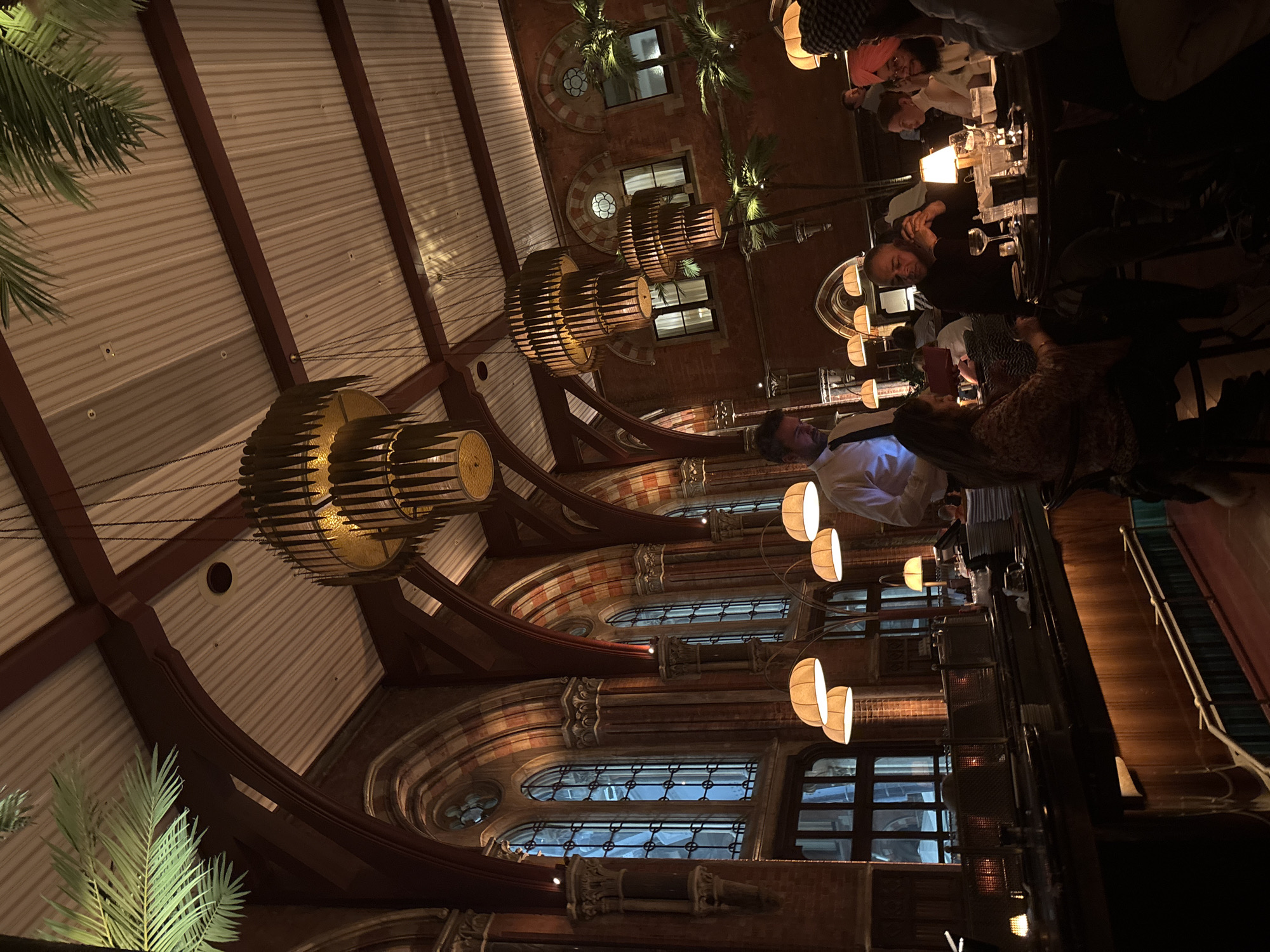 The image depicts the interior of a restaurant, specifically Dishoom King's Cross in London, United Kingdom. The location is characterized by its high ceilings, intricate brickwork, large windows, and decorative lighting fixtures. Patrons are seated at a table, and a staff member is nearby. The general environment is dimly lit.