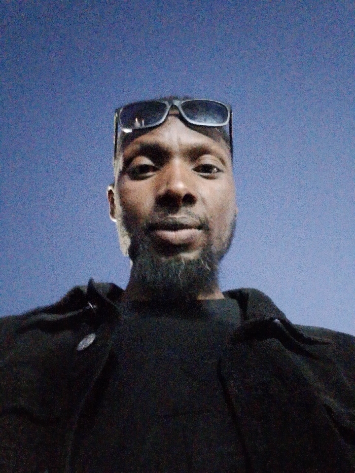 A low-angle selfie of a man with dark skin, a short beard, and sunglasses perched on his head. He wears a dark-colored shirt and jacket. The background is a solid, pale blue, possibly representing a night sky or a blank wall. The lighting suggests an outdoor setting or a location with artificial light sources. The man looks directly at the camera with a neutral expression. The image appears to be taken with a mobile device or similar camera.