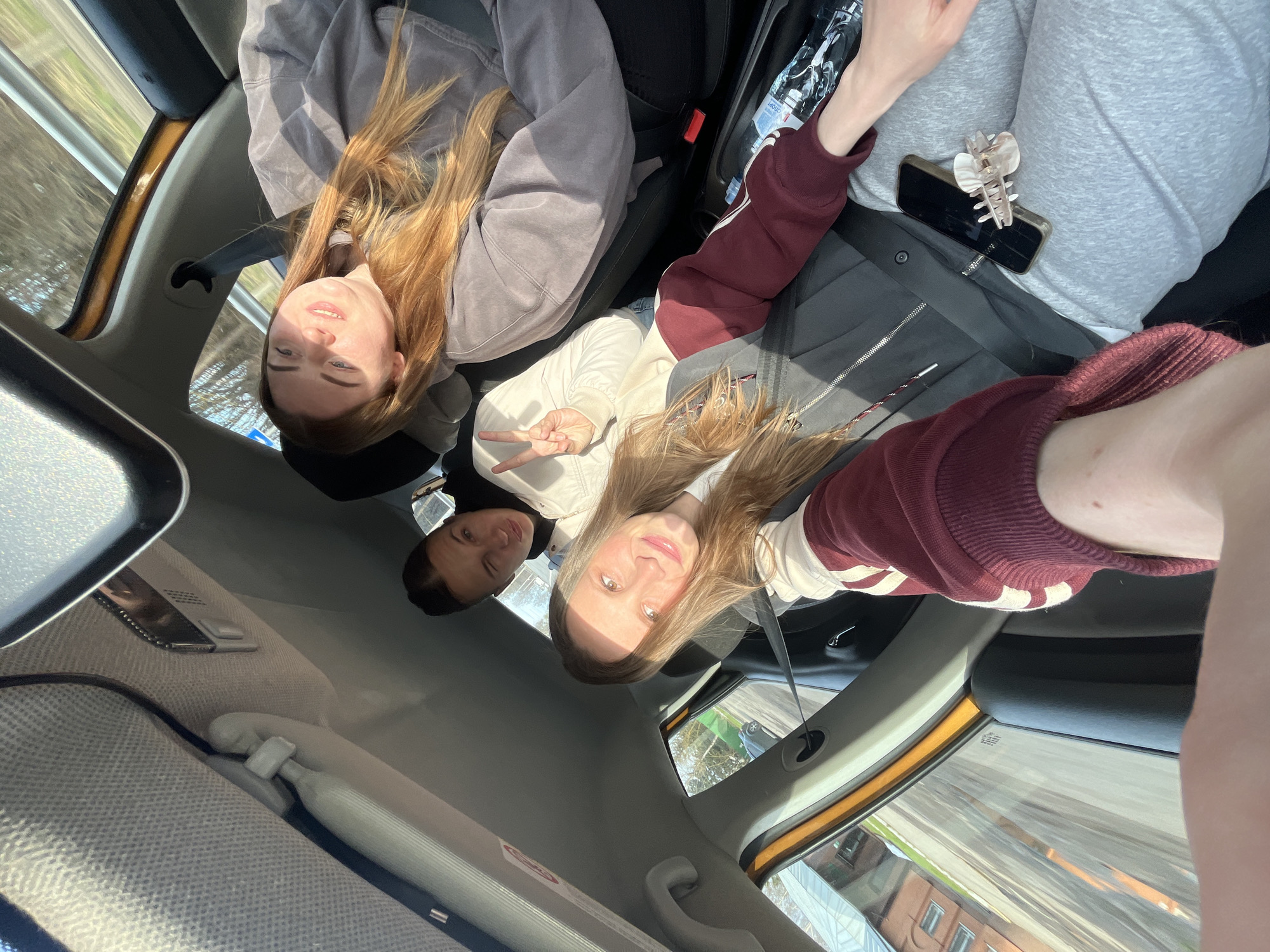 A bright, sunny daytime selfie is captured inside a moving car, featuring four young women. The image is rotated 90 degrees clockwise, offering a high-angle view looking down into the vehicle's interior.
In the foreground, an arm wearing a maroon and white striped long-sleeved top is extended, holding the camera, with a leg in light grey pants also partially visible, belonging to the person taking the photo. Resting on a black smartphone next to this individual is a pearlescent claw hair clip.
Two women are prominently visible in the front seats. The front passenger, central to the frame, has long blonde or light brown hair, wears a white top and a light jacket, and is seen smiling gently while holding up a peace sign with her right hand. Behind the driver's seat, another woman with long reddish-brown hair, clad in a grey-purple hoodie, looks towards the camera with a relaxed expression. A third woman with dark hair and a dark jacket is partially visible in the back seat behind the front passenger, also looking upwards.
The car's interior features dark seats, a light grey headliner, and visible seatbelts. A clear plastic water bottle can be seen near the passenger footwell. Blurred scenery outside the windows, including residential buildings and trees, indicates that the car is in motion under clear, sunny skies. The overall mood is casual, happy, and relaxed, suggesting a shared journey or outing, with the location context being Ширяево, Russia. No discernible text is clearly visible on any objects.