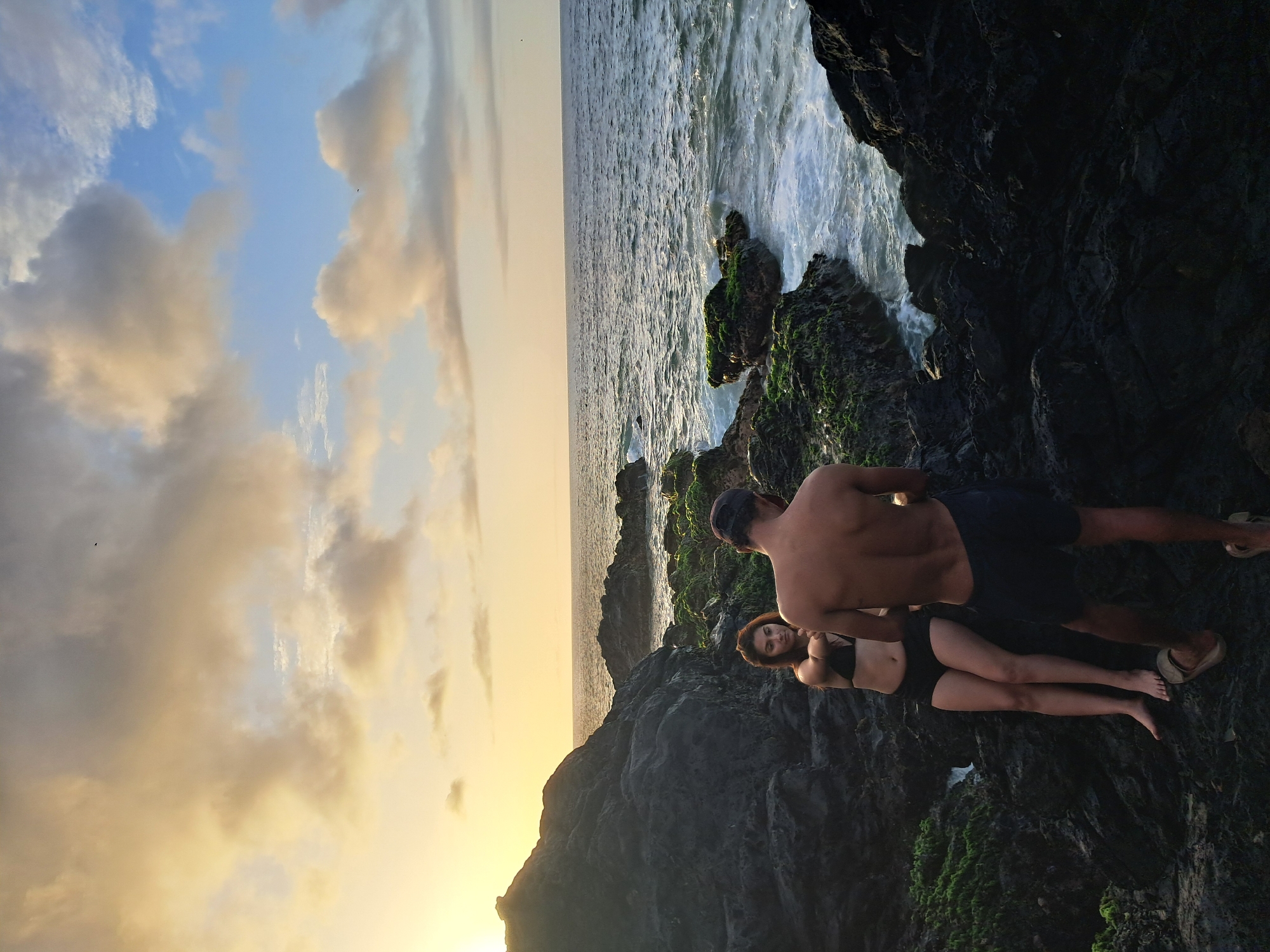 A young couple, a male and a female, stand on a rocky coastline during sunset in Pedro González, Venezuela. The woman, wearing a black bikini, is seated on the rocks, facing the camera with a slight smile. The man, shirtless and wearing dark blue shorts, stands with his back to the camera, looking out at the ocean. The sea is calm with small waves crashing against the moss-covered rocks. The sky is filled with a mix of orange, yellow, and blue clouds, indicating the sun is setting on the horizon. The overall scene is serene and romantic, capturing a moment of natural beauty and connection.