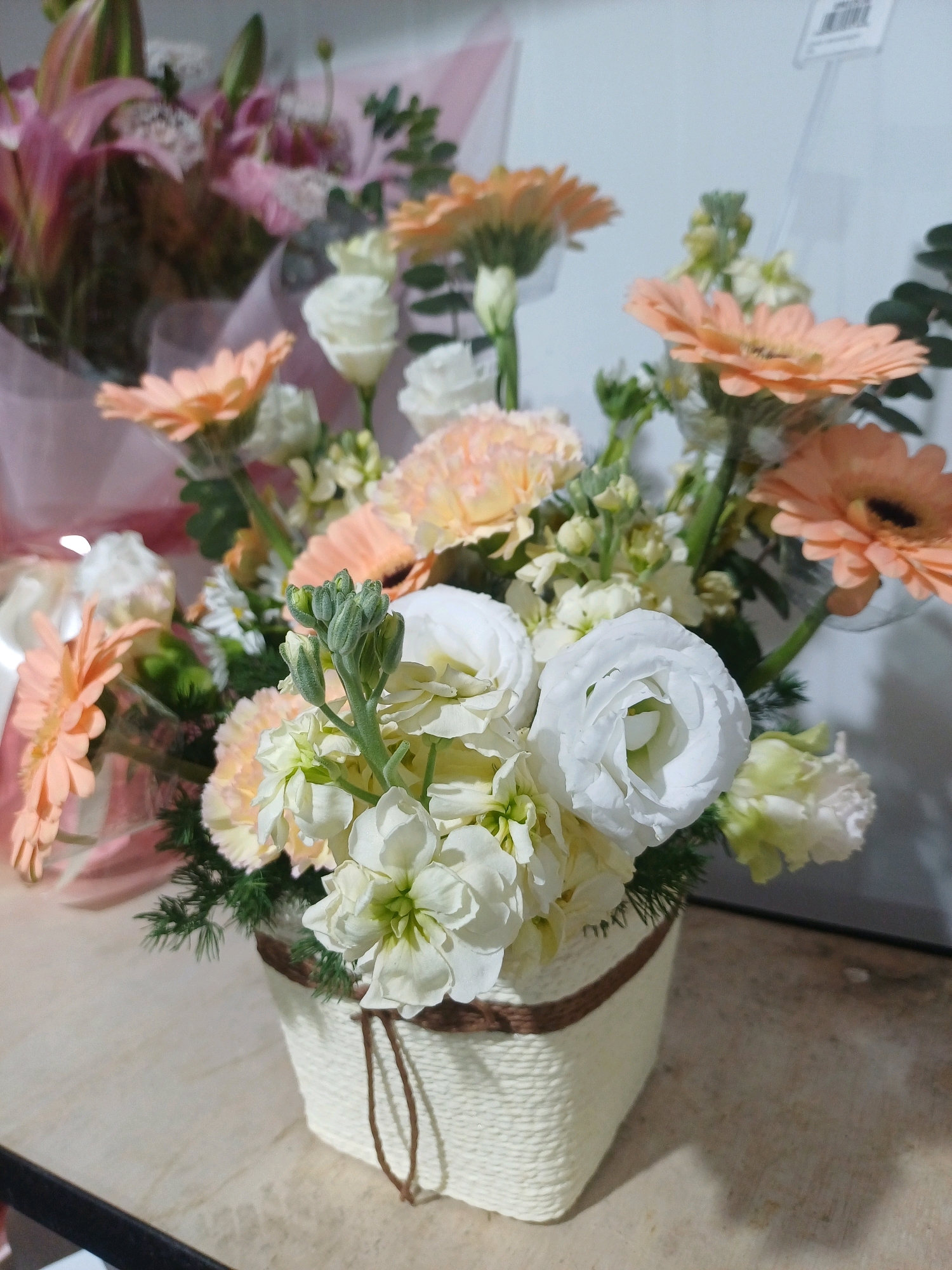 A floral arrangement in a woven basket sits on a textured countertop. The arrangement features peach gerbera daisies, white roses, white carnations, and green foliage. The background is blurred, showing hints of pink lilies and other greenery, suggesting a display or shop environment. The lighting is soft, casting subtle shadows on the countertop. The image is taken from a slightly elevated angle.