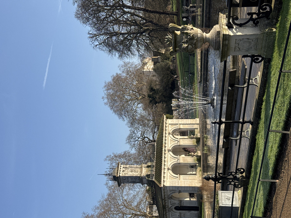 The image displays a sunlit outdoor scene at the Italian Garden in Hyde Park, London, United Kingdom. A classical white stone pavilion, identified as the Pump House, is situated in the mid-ground. It features a dark tiled roof and a prominent, ornate central tower with sculptural details. One individual, dressed in a light pink gown, stands within an archway of the pavilion. Directly in front of the pavilion is a dark pond featuring an active, multi-tiered fountain spraying water.
In the foreground, a black ornate metal railing delineates a grassy strip from a paved area. Two dark park benches are visible behind this railing. To the right, a large decorative stone urn, intricately carved with figures, rests on a stone pedestal. The background consists of a gently sloping grassy area populated by numerous bare deciduous trees. A large, light-colored building is partially visible through the trees in the distance. The sky is clear blue with a single, faint white contrail extending horizontally across the upper portion. Several small, indistinct human figures are present on the distant grassy slope. The scene is illuminated by strong natural sunlight, casting discernible shadows.