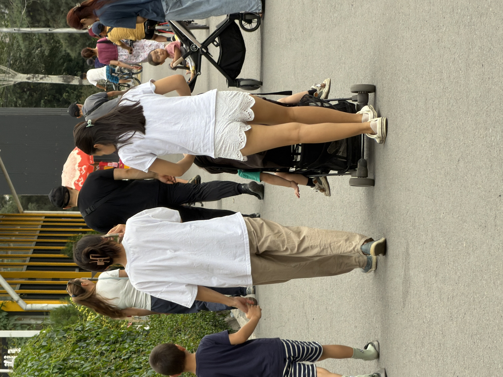 In an outdoor urban setting, a group of people are walking on a paved path, with a lush green hedge on the left and a building with yellow and black vertical accents in the background. In the foreground, a young boy in a navy blue t-shirt and striped shorts holds hands with a woman wearing a white t-shirt and tan pants. Behind them, another woman in a white t-shirt and white lace shorts pushes a stroller containing a child. Further back, several other individuals are visible, including a man in a black t-shirt, a person on a bicycle, and a woman with long brown hair. Some of the people are holding hands or interacting with the children. The scene appears to be a casual outing in a public space.