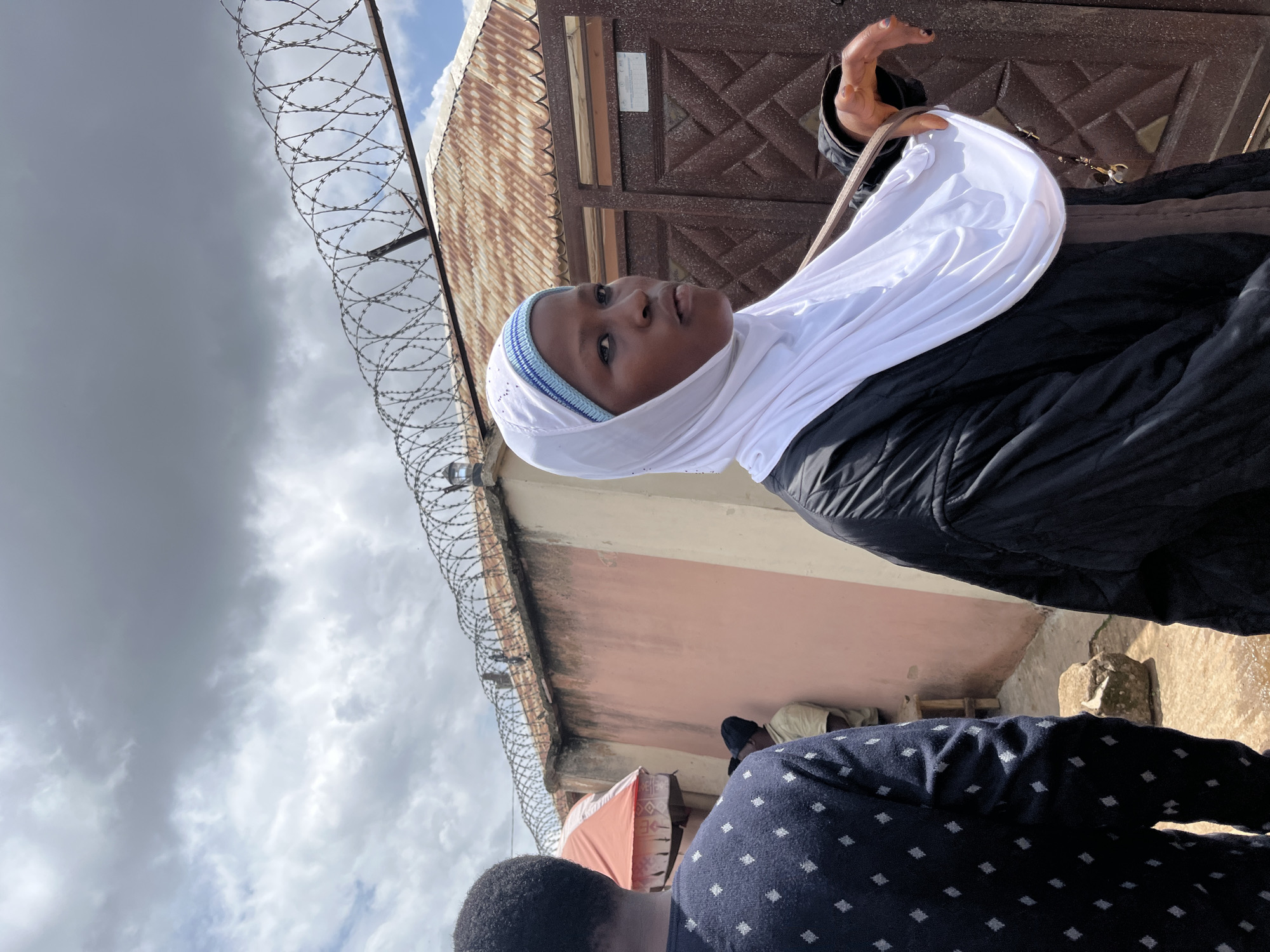 The image captures an outdoor scene in Jos, Nigeria, under a cloudy sky suggesting it is daytime. The foreground is dominated by a young person wearing a white hijab and a black jacket. The individual is looking upwards and to the right with a neutral expression. A hand is visible in the upper right of the frame, near the person's head, with the fingers slightly splayed. Behind the main subject, a corrugated metal roof and a patterned brown gate are visible. Along the top of the image, barbed wire is strung along a metal fence, partially obscuring the sky. The background shows a pinkish-beige wall and hints of other individuals, one wearing a light-colored shirt and a black cap, and another in a dark blue patterned shirt. There is also a glimpse of a small orange awning and some makeshift structures. The ground appears to be rough and possibly wet in some areas. The overall setting suggests a somewhat utilitarian or possibly residential outdoor space, with elements of security fencing. No visible text is present in the image.