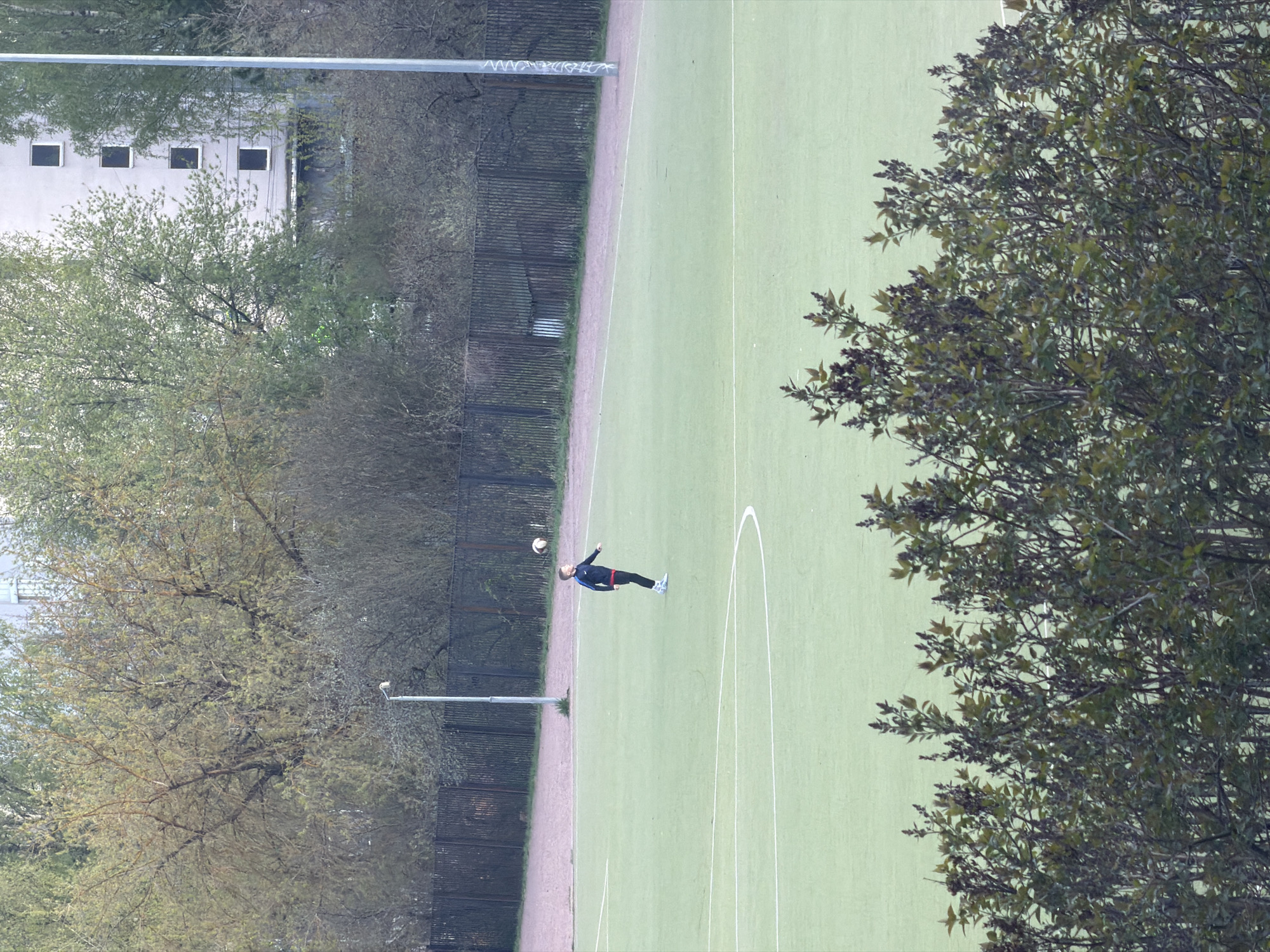 A horizontal aerial shot depicts a soccer field in Myaglovo, Russia. The field is surrounded by trees and a building is visible in the background. A person is kicking a soccer ball on the field.