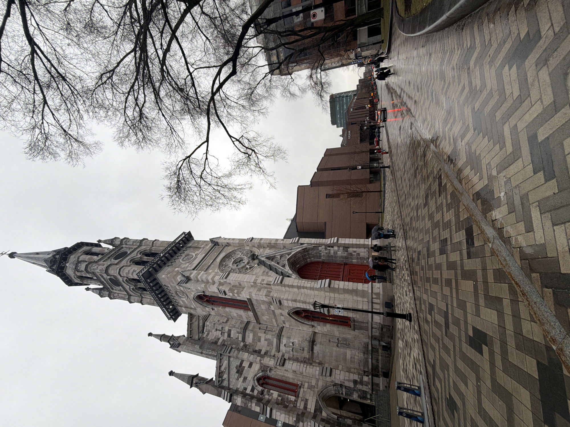 The image shows the Église du Gesù in Montreal, Canada, taken from a low angle on a cloudy day. The church, a prominent Gothic Revival structure, occupies the left side of the frame. A large tree with bare branches is visible above the church. The foreground features a brick-paved walkway, slightly wet, leading towards a street scene with vehicular traffic and pedestrians. Modern buildings are visible along the street, including a building with UQAM lettering. Some individuals are seen with umbrellas on the sidewalk near the church.