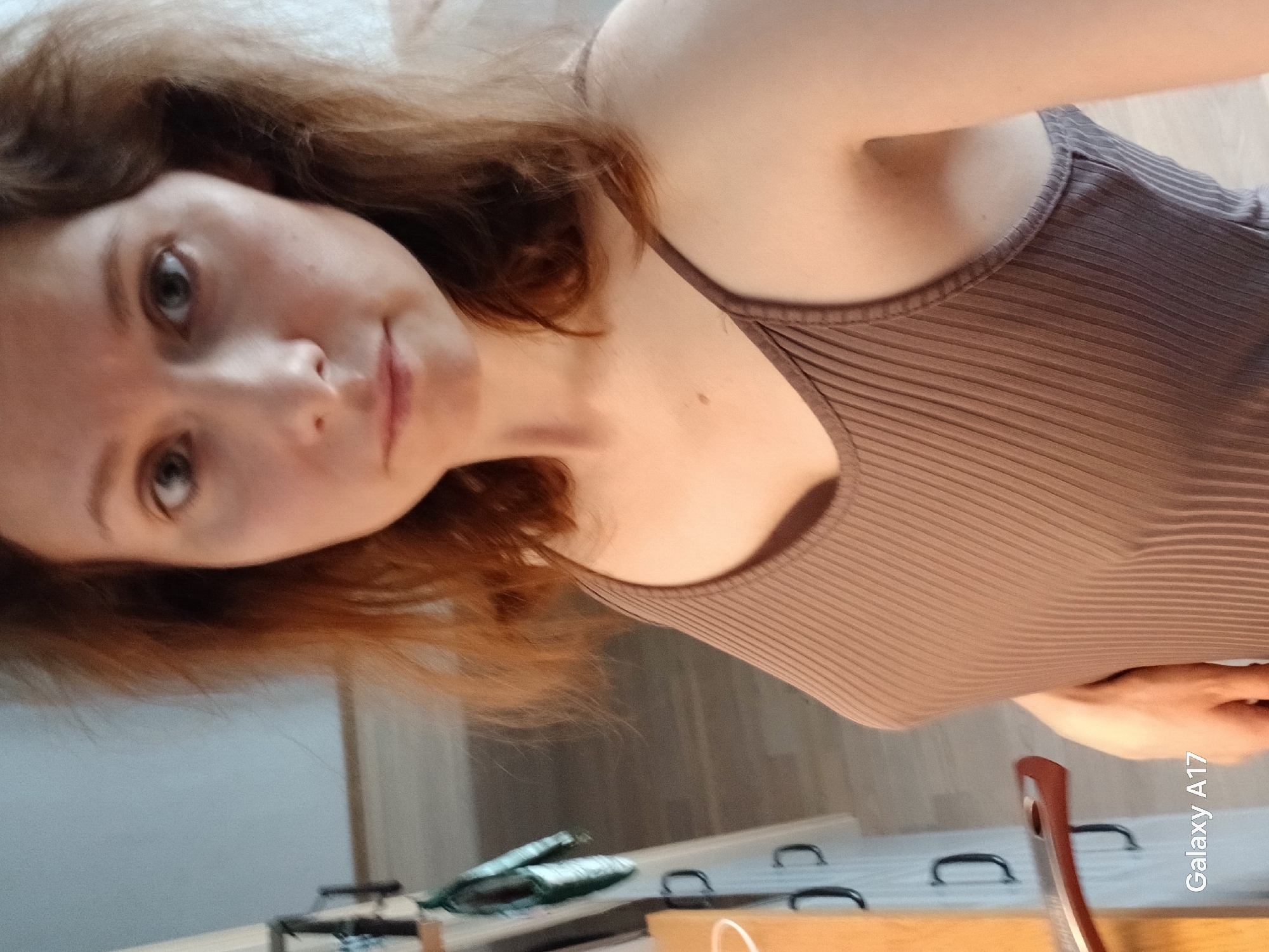 A woman with auburn hair and fair skin is shown in a close-up, angled selfie. She is wearing a brown ribbed tank top. Her gaze is directed towards the camera. The background is blurred, but shows a wooden surface, possibly a dresser or desk, with handles and items that are not clearly discernible. The bottom right corner of the image has the text "Galaxy A17." There is no information present in the image to indicate this photo was taken in Yekaterinburg, Russia.