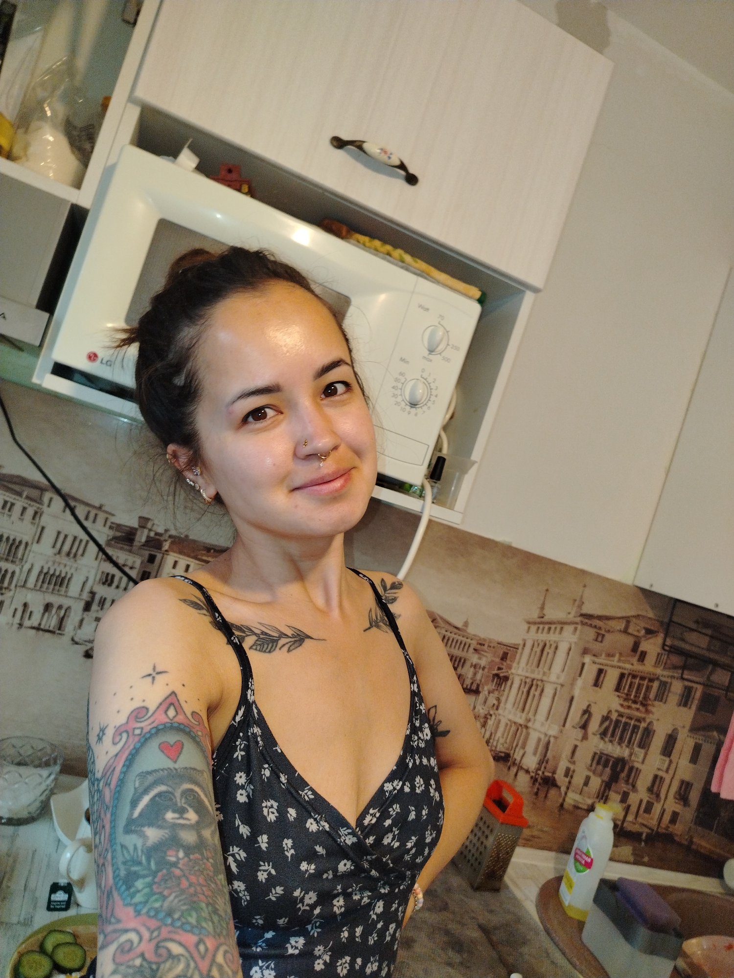 A person with dark hair pulled back, multiple piercings on the left ear, and a nose ring is looking at the camera with a slight smile. The person is wearing a dark-colored dress with a white floral pattern. Visible tattoos are present on both shoulders and the left arm, including a raccoon design.
The background appears to be a kitchen setting. There are white cabinets above, with a white microwave placed underneath. A backsplash with a printed image of a European city, possibly Venice, is visible. Other kitchen items such as a grater, cleaning supplies, and food are scattered on the counter.