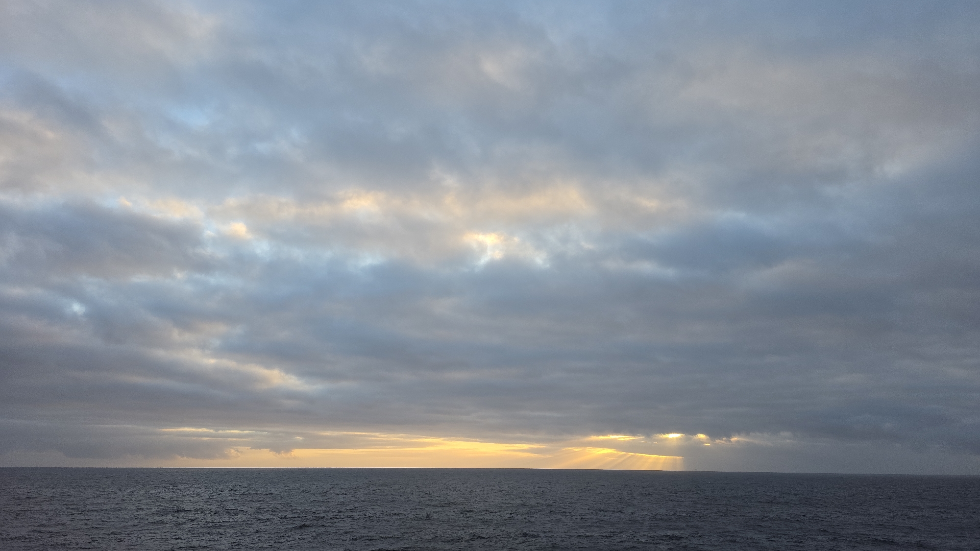 The image depicts a seascape under a cloudy sky at sunset or sunrise. The sky is filled with layers of grey clouds, some with hints of blue and patches where the sunlight is visible, creating a soft golden glow. The horizon is a straight line where the sky meets the dark, rippling water. Several sunbeams are seen extending from the horizon toward the sea. There are no visible people or structures. The location is at sea; the exact location is not identifiable.