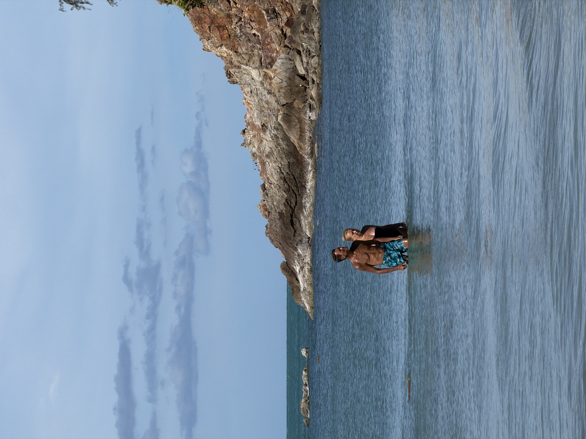 A man and a woman stand in shallow, blue water near a rocky cliff face. The man, in patterned blue swim trunks, faces left with his head turned towards the woman. He has a muscular build and appears tanned. The woman, wearing a black top, stands to his left and slightly behind him, facing him. Her hair is light-colored.
The scene is set against a backdrop of a rugged, brown and grey rock formation that rises steeply from the water's edge. The sky above is a pale blue with wispy, grey clouds. The water is calm with gentle ripples. In the distance, a faint line of rocks juts out of the sea. The image appears to be taken during daylight hours, likely mid-morning or afternoon, given the bright sky and sunlit rocks. The overall mood is serene and relaxed. The location is identified as Baan Plai Laem, Thailand. There is no visible text in the image.