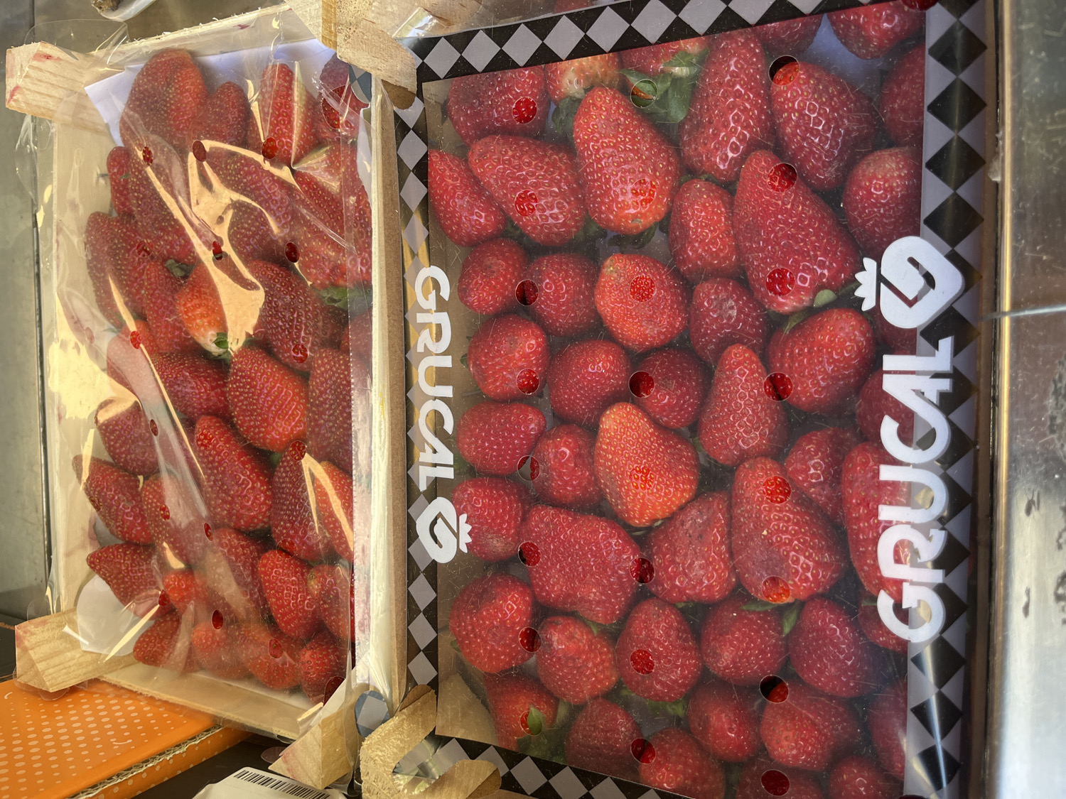Two wooden crates filled with fresh red strawberries are displayed, likely in a market or commercial setting. The image is taken from a slightly elevated, oblique angle, looking down into the crates.
The crate on the left contains a generous quantity of vibrant red strawberries, each appearing plump and ripe. It is covered with a clear plastic film, sealed around the top, with numerous small, circular red stickers or dots affixed to the plastic, some directly over the fruit. The crate itself is made of light-colored wood, showing a simple, functional construction. A white paper or fabric layer is visible beneath some of the strawberries. Below this crate, a textured orange surface and a small white label with faint black text are visible.
The crate on the right, positioned slightly behind and to the right of the first, also holds numerous red strawberries. This crate features more distinct packaging; its upper rim is adorned with a black and white checkerboard pattern. A prominent white logo and the brand name "GRUCAL" are printed vertically on the right side of the plastic film covering this crate. This film also has a few scattered red circular markings, similar to those on the left crate, though fewer in number. The strawberries in this crate also appear fresh and of good quality.
No people are visible in the frame. The overall impression is one of fresh produce being presented for sale or transport, characteristic of a daytime market or grocery display. The branding on the right crate suggests commercial packaging and distribution.