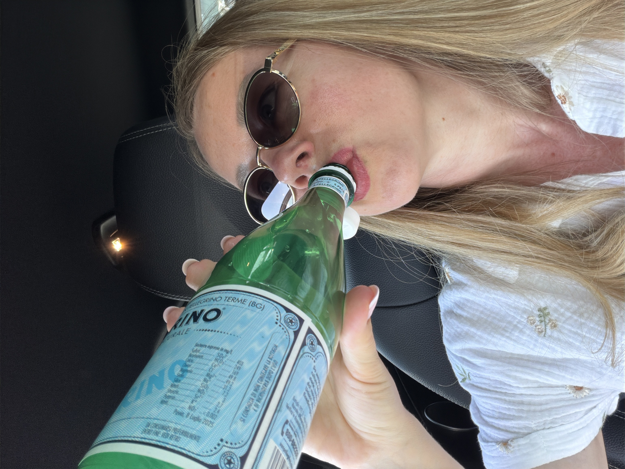 A woman with blonde hair and sunglasses drinks from a green glass bottle of San Pellegrino water in a car. The bottle has a blue and white label with Italian text. The woman is wearing a white top with embroidered flowers. The interior of the car is visible, including a black leather seat and a small light on the ceiling.