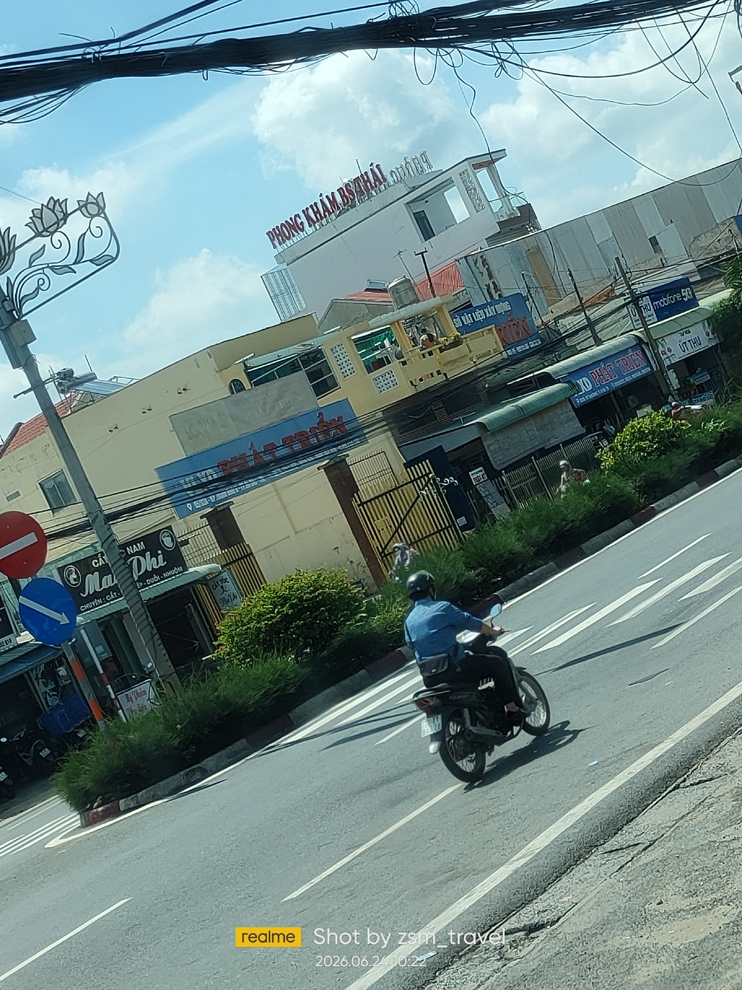 The image captures a street scene in Gò Dầu, Vietnam, on a bright, overcast day. The camera angle is tilted, looking down from a moving vehicle, likely a car or bus.
In the foreground, a person wearing a blue shirt and a black helmet is riding a dark-colored motorcycle down the right side of a paved road. The motorcycle has a license plate that reads "62 B2 - 28". White lane markings are visible on the road. The rider's right leg is bent, and their left leg is positioned over the motorcycle's frame. A black bag is slung across their back.
To the left of the road, there is a sidewalk area with a metal pole supporting traffic signs. One sign is circular with a red border and a horizontal white bar, indicating a prohibition. Below it is a blue circular sign with a white arrow pointing to the left. Next to the pole is a small shop or stall with a blue awning. Further left, a section of green foliage is visible, and behind it, a tall, yellow building with a red-tiled roof. Another building, a clinic identified by the sign "PHÒNG KHÁM BS THÁI" in red lettering, is visible further back, with a modern architectural style and large windows.
The background features a mix of buildings, many with light-colored facades and some with corrugated metal siding. Signs in Vietnamese are present on various buildings, including "sở vật liệu xây dựng" (construction materials) and "mobilefon 5G". Overhead, a dense network of black electrical wires crisscrosses the sky. The sky itself is bright blue with scattered white clouds.
The overall impression is one of a busy urban environment with a blend of commercial establishments and residential structures. The image conveys a sense of everyday life in Gò Dầu.