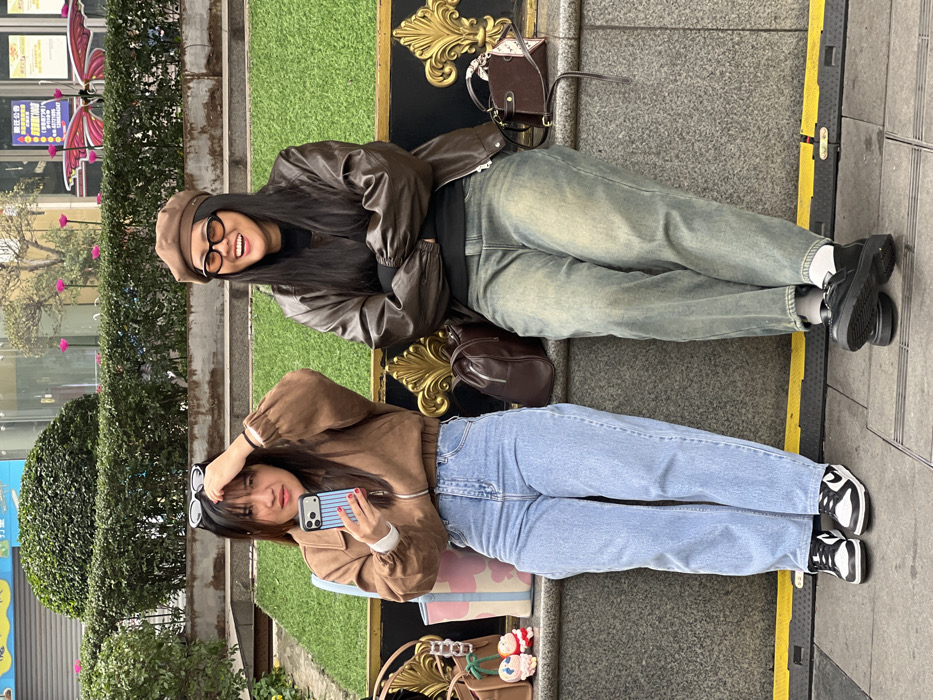 Two young women are sitting side by side on a bench in an urban area. The woman on top wears a brown beret, glasses, a brown jacket, and faded blue jeans. The woman below has sunglasses on her forehead, a brown cropped jacket, light blue jeans, and is holding a phone. Each woman has a purse, and they both wear sneakers. Green hedges and trees are in the background.