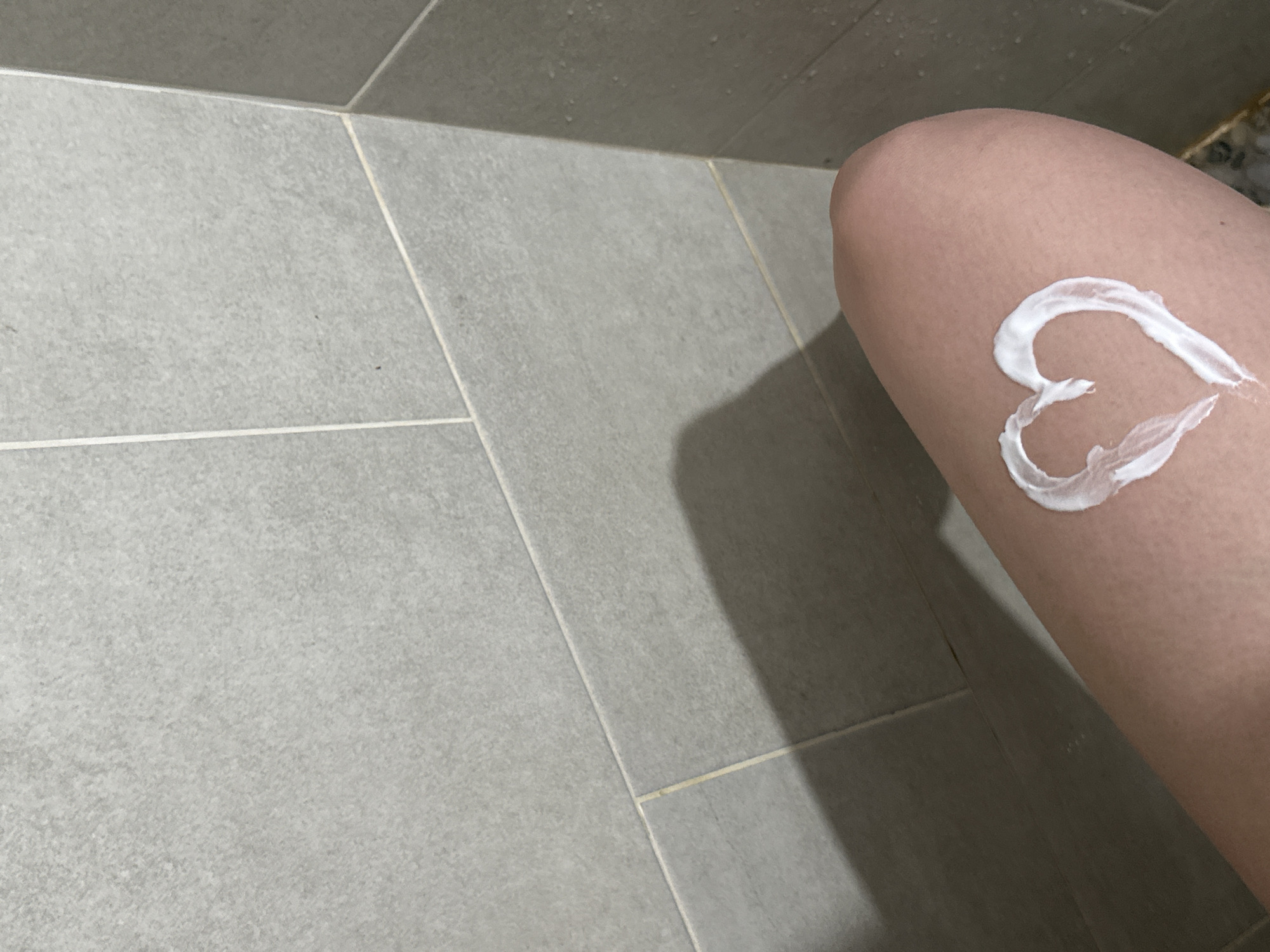 A close-up shot depicts a person's thigh with a heart-shaped dollop of white cream or lotion applied to it. The background consists of grey ceramic tiles with grout lines. The location is not evident from the image.
