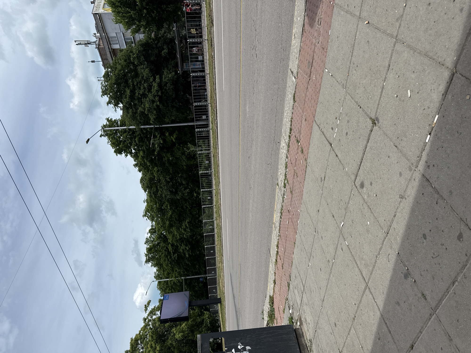 The image shows a street scene with a sidewalk in the foreground and a road in the middle ground. On the left side of the sidewalk, there is a black trash can. The sidewalk is made of concrete tiles and has a section of red brick pavers. There are cigarette butts scattered on the sidewalk. A road with yellow dividing lines runs horizontally across the image. Beyond the road, there is a metal fence, a line of trees, and some buildings. A large digital billboard is visible on the left, and a bus stop shelter with people is visible further down the road on the right. The sky is overcast with clouds.
