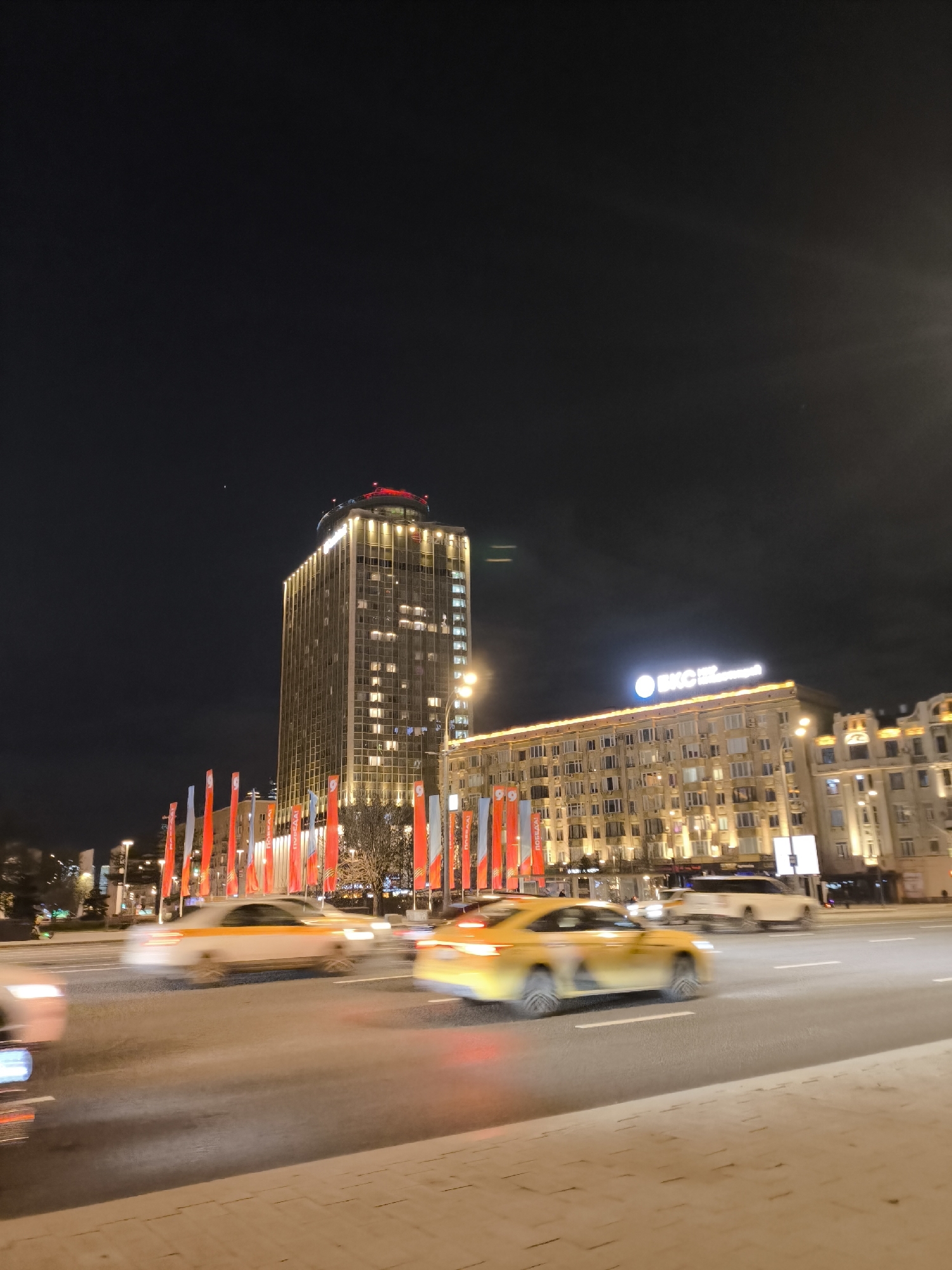 This night-time photograph captures a bustling street scene in Moscow, Russia. The foreground features a multi-lane road where several vehicles, including yellow taxis, are blurred due to motion, indicating active traffic.
Two prominent buildings anchor the background. On the left stands a tall, modern skyscraper with numerous illuminated windows, topped by a distinctive red-lit structure. To its right, a lower, classical-style building is brightly lit, showcasing its architectural details. This building also features a prominent illuminated sign displaying the Cyrillic text "БКС" (BKS) with smaller letters below.
The street is adorned with numerous red and white vertical banners or flags, suggesting a celebratory or commemorative occasion. On some of these banners, the Cyrillic word "ПОБЕДА!" (Victory!) is discernible, along with the number "99", strongly linking the scene to Victory Day celebrations.
The time of day is night, evidenced by the dark sky and the extensive artificial illumination from buildings, streetlights, and vehicle lights. The sky appears clear, without adverse weather. No individuals are clearly visible in the frame, with the focus primarily on the urban landscape, architecture, and traffic. The overall impression is one of a vibrant and active city street.