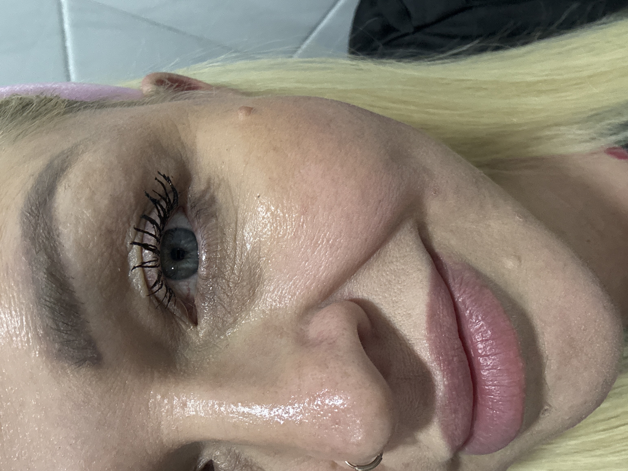 The image is a close-up, eye-level, medium shot of a Caucasian woman's face, focusing on her right eye, nose, and lips. She has blue eyes, blonde hair, and is wearing dark mascara. A pink headband is visible on her head. Her skin appears shiny, possibly due to sweat or makeup. A small mole is visible on her right cheek. A silver hoop earring is in her left nostril. The background is out of focus and appears to be a tiled wall, possibly in a bathroom. No specific location in Batumi, Georgia is indicated by the image.
