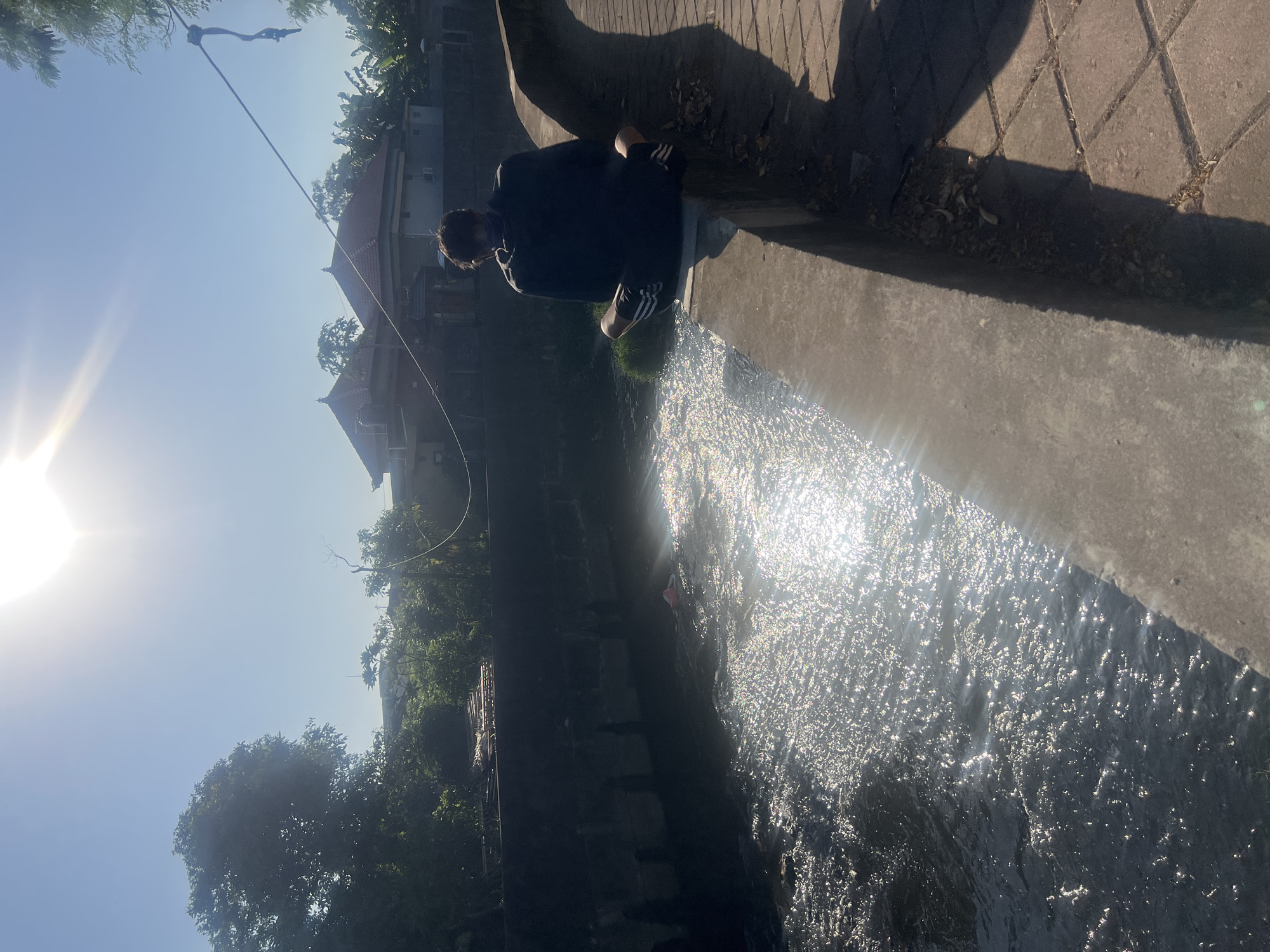 A person sits on the edge of a concrete embankment next to a narrow, murky canal filled with glittering sunlight. The person, viewed from behind, wears a dark hooded sweatshirt and black shorts with white stripes. Their legs are crossed, and they are facing away from the camera, looking towards the canal and the buildings beyond. The canal has a dark, artificial-looking wall on the left, with some debris visible in the water. On the right side of the canal is a paved walkway. In the background, across the canal, are several buildings with traditional Indonesian-style roofs, surrounded by lush green trees. The sky is clear and bright blue, with the sun high and shining intensely, creating lens flare. A thin rope is strung across the upper right of the frame, attached to a pole. The scene appears to be in a semi-urban or residential area of Denpasar, Indonesia, possibly a drainage canal or a small river running through a neighborhood.