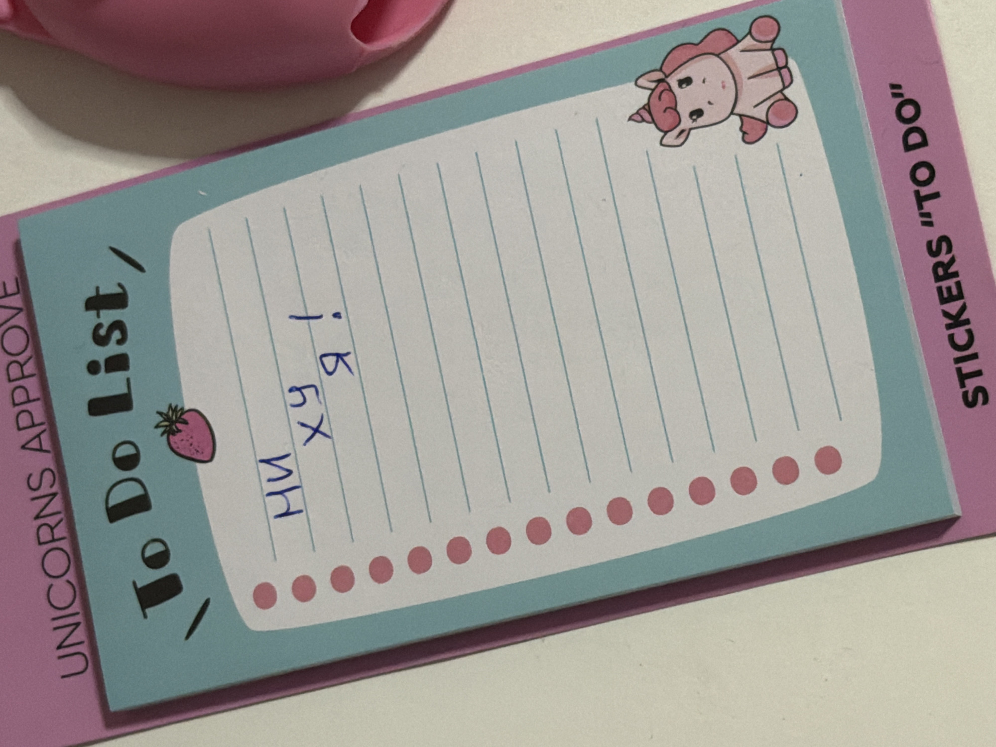 The image shows a close-up of a pink and blue to-do list notepad with a drawing of a unicorn on it. The notepad has lines for writing and several pink dots down the left side. There is blue handwriting on the notepad that says "НИ ху я!". At the top of the notepad, it says "UNICORNS APPROVE" and "To Do List," and at the bottom, it says "STICKERS "TO DO"". The background is a plain white surface, and a portion of a pink object is visible on the right side of the image. There is no indication of the city of Moscow, Russia, or any people present in the image.
