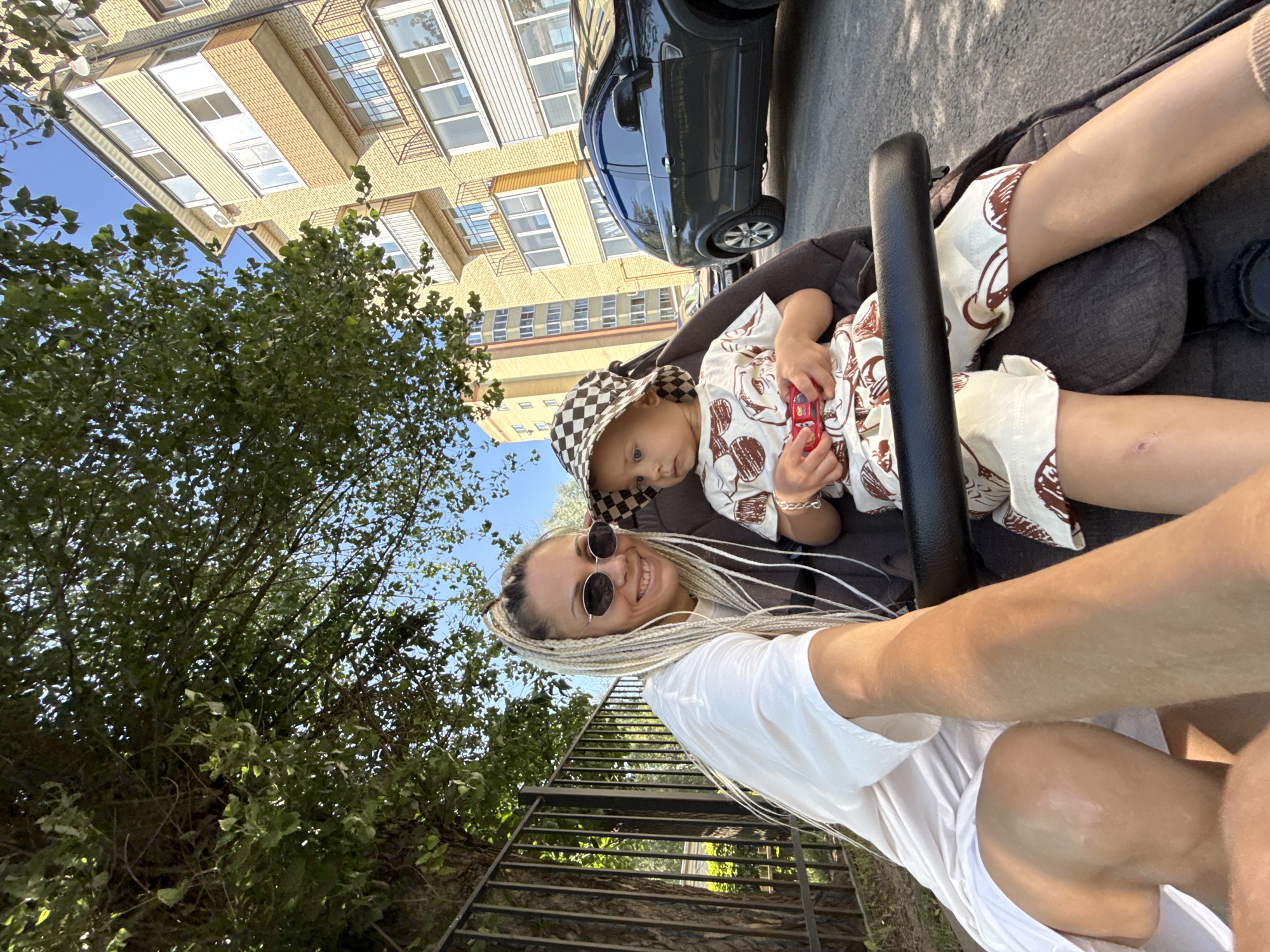 A woman and a child are in a stroller in front of a building. The woman is wearing sunglasses and a white shirt. The child is wearing a checkered hat and a patterned outfit. The child is holding a toy car. The background shows a building with balconies and a car parked in front of it. The scene is outdoors on a sunny day.