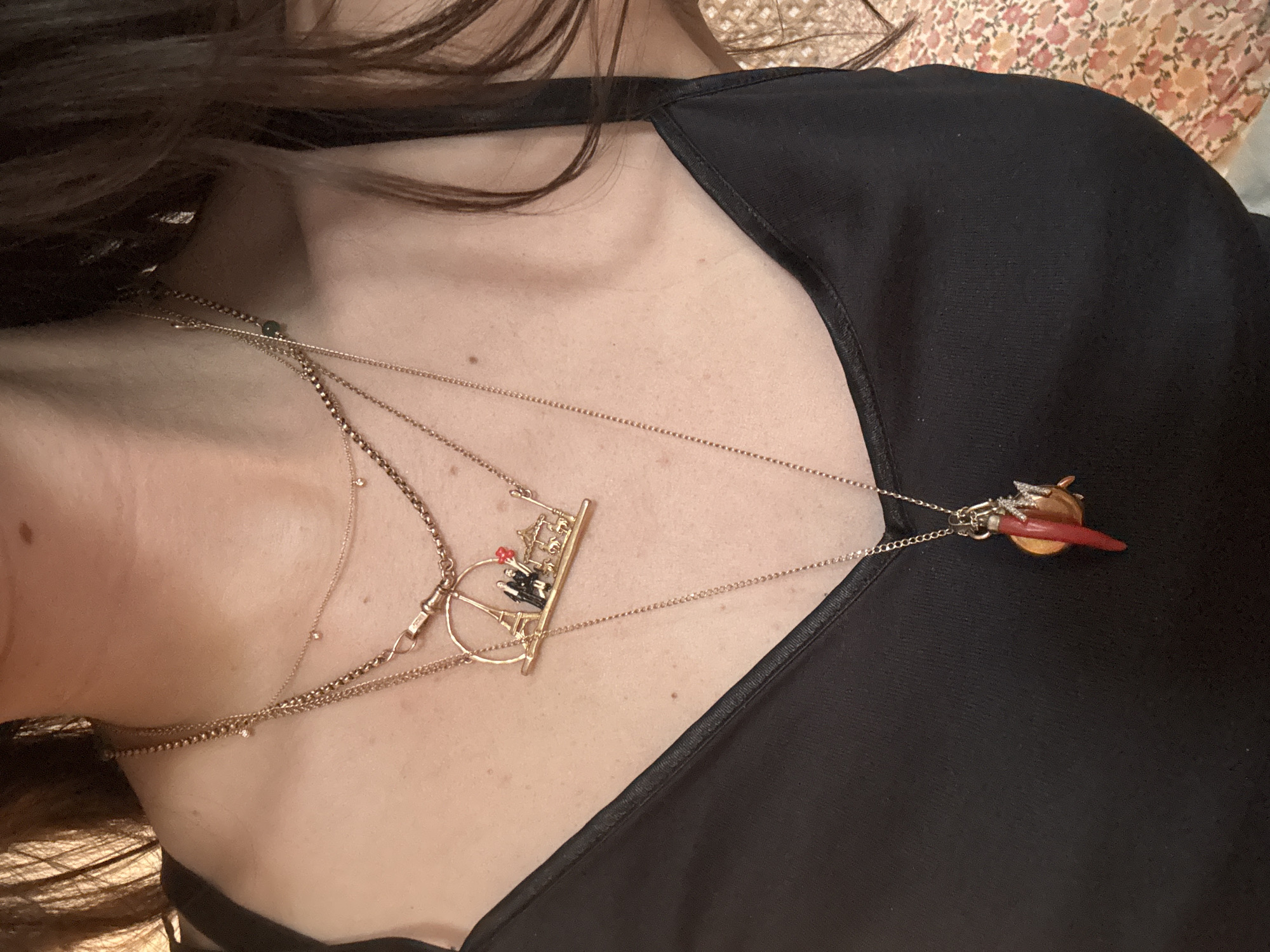 A woman wears several gold necklaces, one with a charm depicting a street scene from Paris, France, including the Eiffel Tower. Another necklace features a horn-shaped charm of an orange color. The woman wears a black top with a v-neck. Her hair is dark brown. The image is a close-up view of the woman's chest and neck.