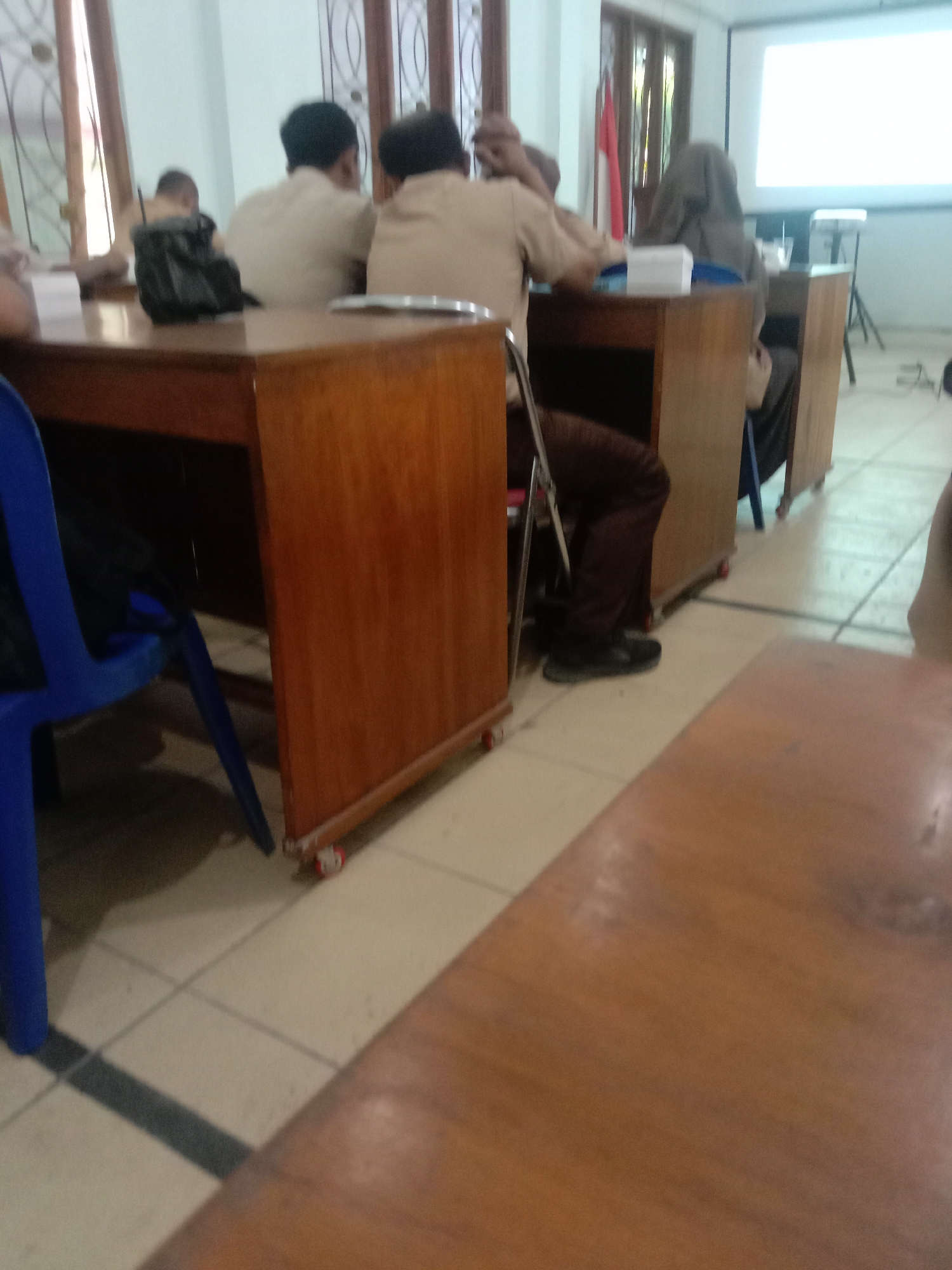 The image depicts an indoor scene, likely a classroom or meeting room, with several individuals present. In the foreground, a light-colored tiled floor is visible, with dark grout lines forming a grid pattern. A wooden table, with a blurred surface, occupies the lower right portion of the frame.
In the mid-ground, a blue plastic chair is positioned on the left, partially obscuring a wooden desk. Behind this desk, two individuals wearing light-colored, possibly uniform, tops are seated, facing away from the camera. One of them has a black bag placed on the desk. Further back, another person, wearing a dark head covering, is seated at a desk. To the left of this person, a red and white Indonesian flag is visible, partially obscured by a wooden doorframe. In the background, a projection screen is illuminated with a white light, suggesting a presentation is in progress. The overall lighting appears to be from overhead sources. The location is identified as Kalisari, Indonesia.