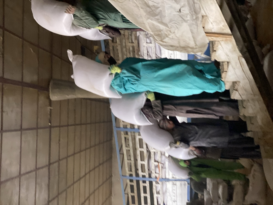 People wearing green and blue coveralls carry white sacks through a warehouse. The warehouse has a metal grid ceiling and metal support beams. Some people appear to be wearing gloves. A white tarp is visible in the background. The sacks are large and heavy. The scene suggests a storage or distribution facility.