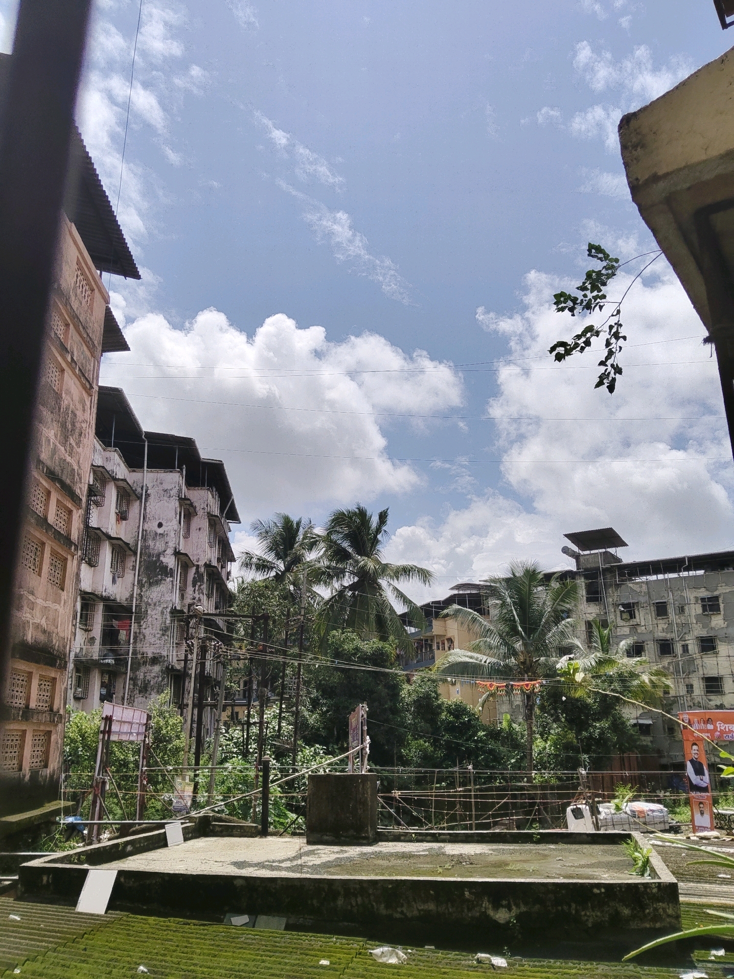The image captures a view of buildings and trees under a partly cloudy sky, likely in Vasai-Virar, India. In the foreground, a rooftop area with a concrete structure and some green mossy surfaces is visible. Beyond this, several apartment buildings with multiple stories and balconies stand. Lush green trees, including palm trees, are interspersed between the buildings, suggesting a tropical or subtropical climate. Electrical wires and poles are also present, indicating an urban or semi-urban setting. On the right side, a large orange banner with images of people is partially visible. The overall scene suggests a residential area with a mix of construction and natural elements.