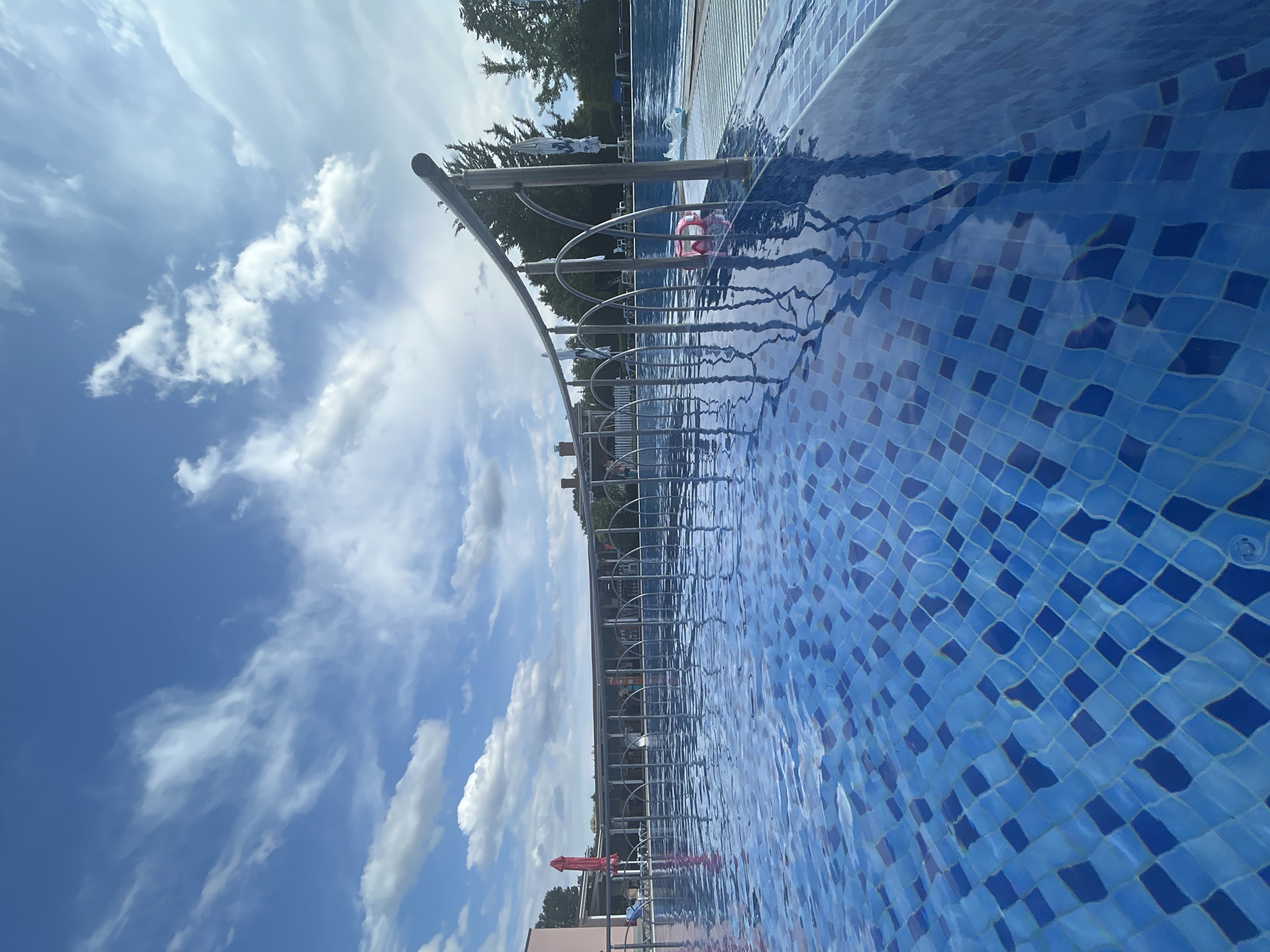 The image shows an outdoor swimming pool with blue tiled walls. A metal railing with vertical supports curves along the edge of the pool. The water is clear and reflects the sky, which is partly cloudy with scattered white clouds against a bright blue sky. In the background, there is a building with a pink facade and red umbrellas. Trees and more swimming pool structures are visible in the distance. There are no people clearly visible in the foreground or engaged in any activity. The location is described as Unknown, Unknown.
