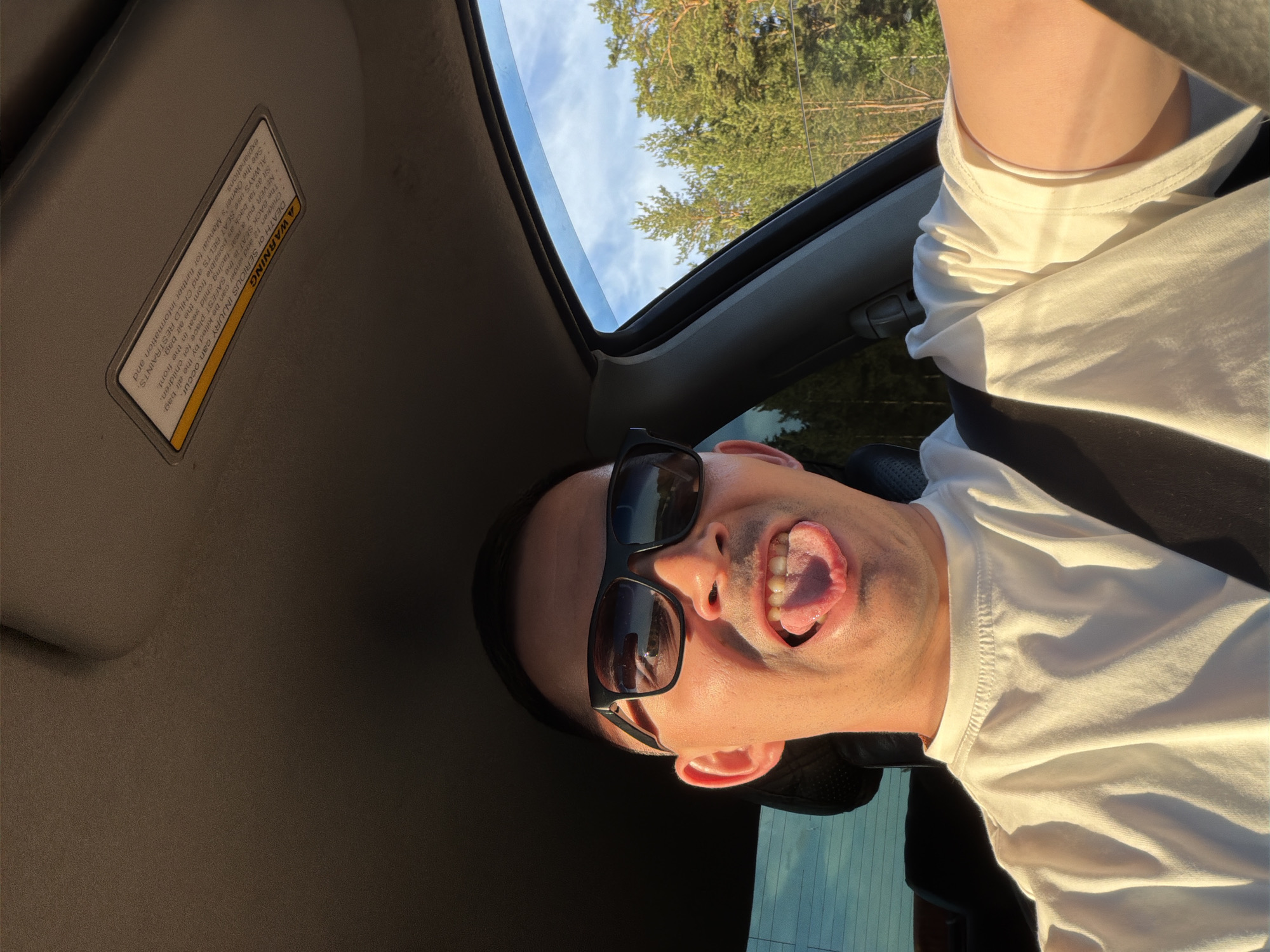 A playfully posed portrait features a man from the chest up, positioned inside a car and looking directly at the camera. He wears dark sunglasses and a white t-shirt. His head is tilted, and he has a humorous expression with his mouth open and tongue slightly extended. Warm, golden sunlight illuminates his face and clothing, suggesting the late afternoon or early evening.
Above the man, the open sunroof of the car reveals a clear blue sky with some scattered clouds, and the lush green tops of coniferous trees, indicating an outdoor setting, likely in a natural or forested area. The interior of the car, including the tan headliner and a sun visor, is visible.
A clear warning label is affixed to the car's sun visor. The text on the label states: "A WARNING DEATH OF SERIOUS INJURY can occur. Children 12 and under can be killed by the air bag. The BACK SEAT is the SAFEST place for children. NEVER put a rear-facing child seat in the front. Sit as far back as possible from the air bag. ALWAYS use SEAT BELTS and CHILD RESTRAINTS. See the Owner's Manual for further information and explanations." The image captures a lighthearted moment, possibly during a car journey in Yus'ki, Russia.