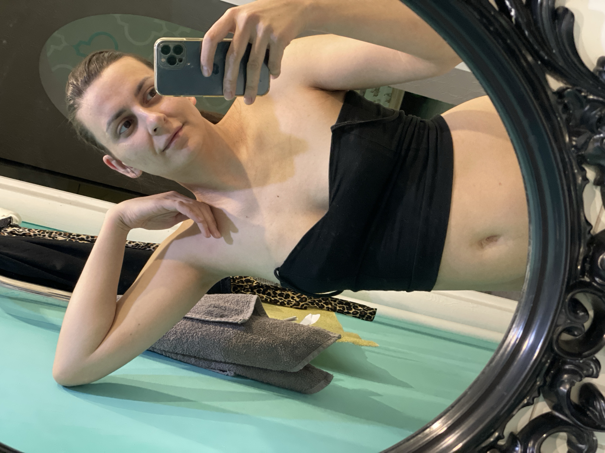 A selfie in a black ornate mirror shows a woman in a black top lying on her side with one arm propped under her head and the other holding a phone. A gray towel is stacked beside her, with a patterned scarf partially visible underneath. The surface beneath is a light blue or green. The location appears to be indoors.