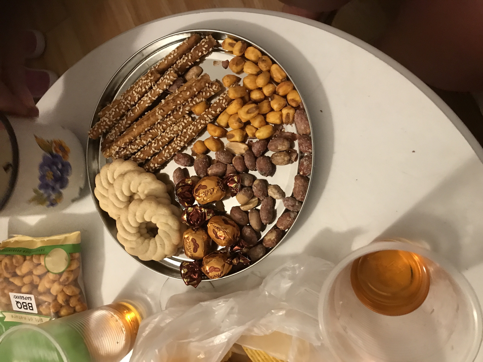 The image depicts a close-up overhead view of a white table surface, possibly in a residential setting in Varna, Bulgaria. A silver platter is centrally positioned, laden with various snacks. These include two round, light-colored cookies with a ridged texture, a row of thin, sesame-seed-covered breadsticks, and a variety of nuts, some appearing to be peanuts with reddish skins and others golden-yellow. Several individually wrapped chocolates with gold and red foil wrappers, some labeled "Coffee," are also present on the platter. To the left of the platter, a clear plastic cup filled with an amber-colored liquid, likely beer or a similar beverage, is visible. Behind this cup, a portion of a partially opened bag of snacks, featuring text that includes "BBQ," is visible. The background is slightly blurred, showing hints of other objects and surfaces. The lighting suggests an indoor environment, possibly an evening setting.