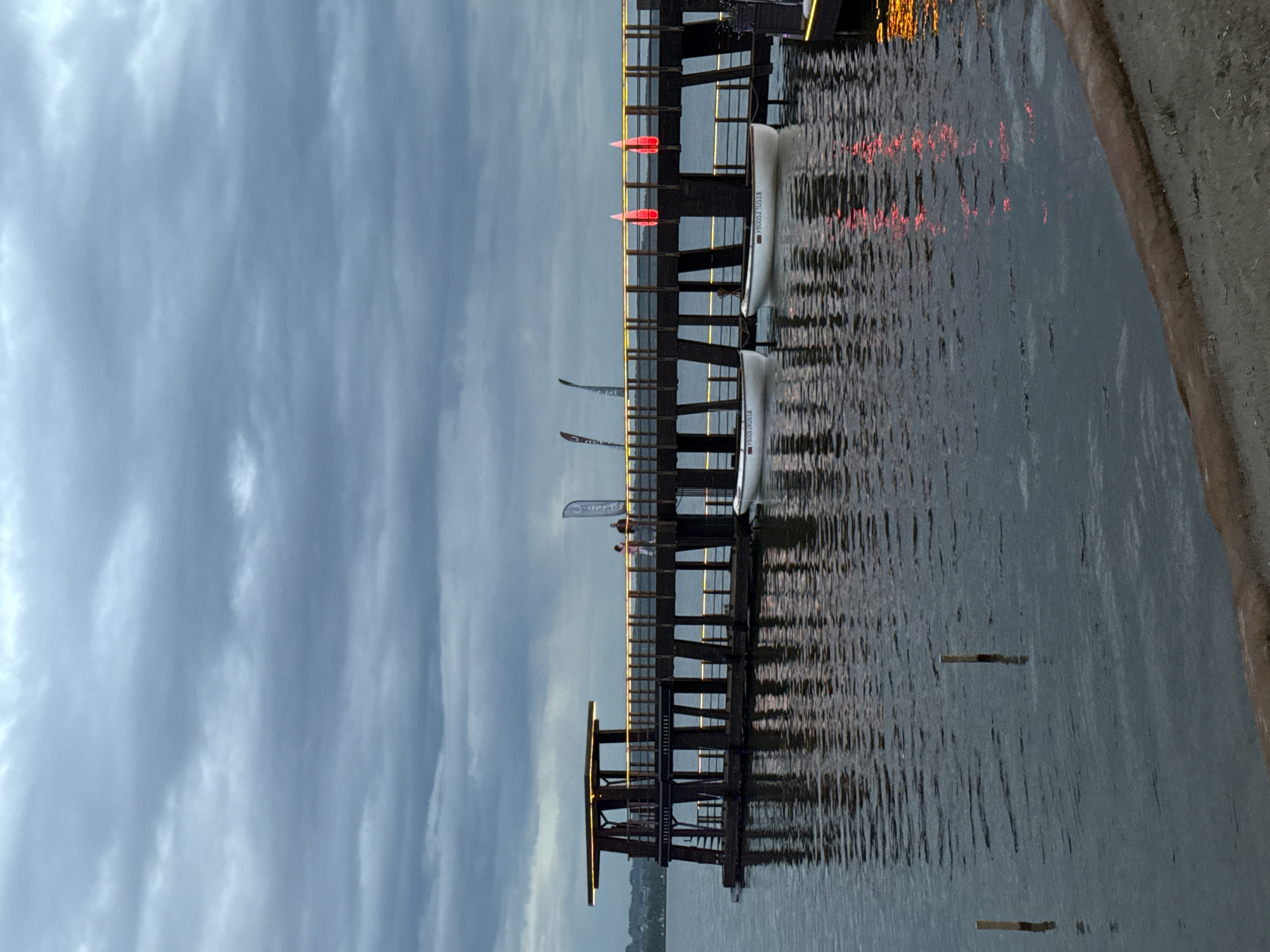 An outdoor scene depicts a wooden pier extending over dark, rippling water under a cloudy, overcast sky. The pier is supported by numerous vertical posts and features a raised, covered structure on the left end with a flat roof illuminated by yellow lights. Along the edge of the pier, yellow lights are visible, and two red, illuminated objects, possibly lights or flags, are spaced apart. Two white rowboats are docked on the water, one with visible markings that read "Y8005TRUS18" and "Y80052US18". Two individuals, a man and a woman, are walking on the pier, and a dark flag with a white logo is attached to a pole on the right side of the pier. The water reflects the pier's lights and structure. The scene appears to be in Votkinsk, Russia.