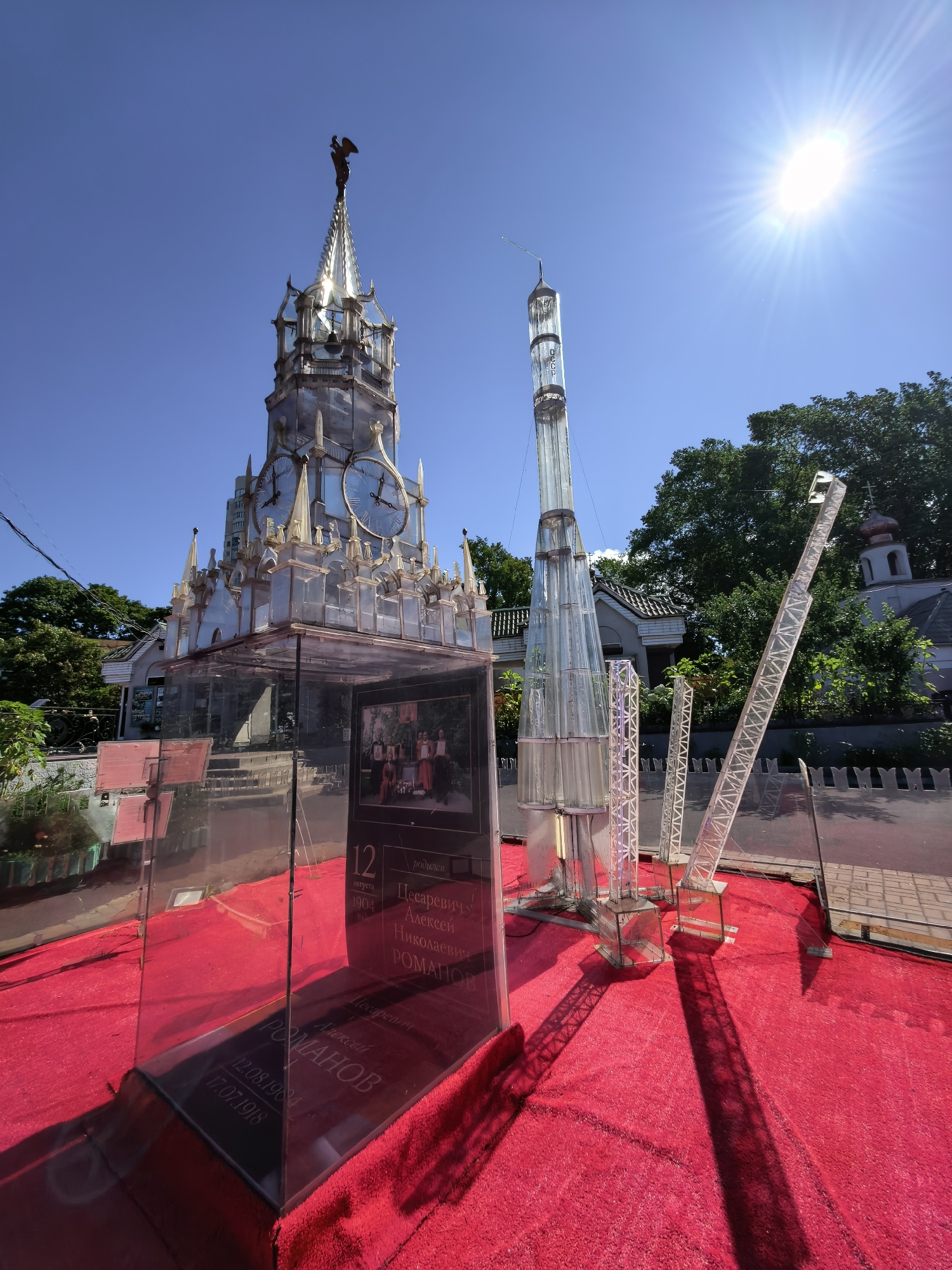 This image captures an outdoor exhibition on a bright, sunny day in Sestroretsk, Russia. In the foreground, a vibrant red carpet covers the ground, leading to a transparent display case containing a dark plaque with engraved text and a framed photograph of a group of people. To the right of the display case, a tall, glass-like model resembling a rocket stands prominently. Behind it, a detailed model of a clock tower, also appearing to be constructed from glass-like materials, dominates the left side of the frame, topped with a figure and featuring large clock faces. Several metal scaffolding structures, possibly part of the exhibit, are also visible. In the background, lush green trees and traditional Russian architecture, including a building with a copper-domed roof and a cross, are present. The sun shines brightly in the upper right corner, casting long shadows across the scene. The overall impression is of a public art installation or historical exhibit presented outdoors.