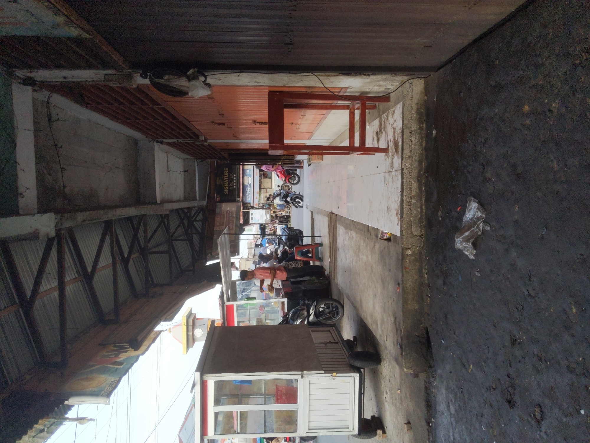 The image depicts a street scene in Keudematangpanyang, Indonesia, likely during daylight hours. The foreground is dominated by a dark, somewhat muddy ground surface, with scattered debris including plastic. A raised concrete curb separates this area from a tiled walkway.
On the walkway, a white and red food cart with glass display cases and wheels is positioned. To the right of this cart, another, similar food cart is visible, attended by a man in a pink shirt. He appears to be serving or preparing food, with several motorcycles parked nearby. One motorcycle, a black scooter, is prominently placed in front of the first food cart, and another red motorcycle is behind the second food cart. A person is seated on a red stool near the second food cart.
Further down the walkway, a tall, dark wooden structure, possibly a stall or shop, runs along the right side of the image. It features a corrugated metal roof supported by a dark metal frame. A sign with the words "SIGNATURE KRETEK" and five stars is visible on the wall of one of the buildings. In the background, a clock tower with a yellow and red spire can be seen. The overall environment suggests a commercial or market area with an outdoor food vendors.